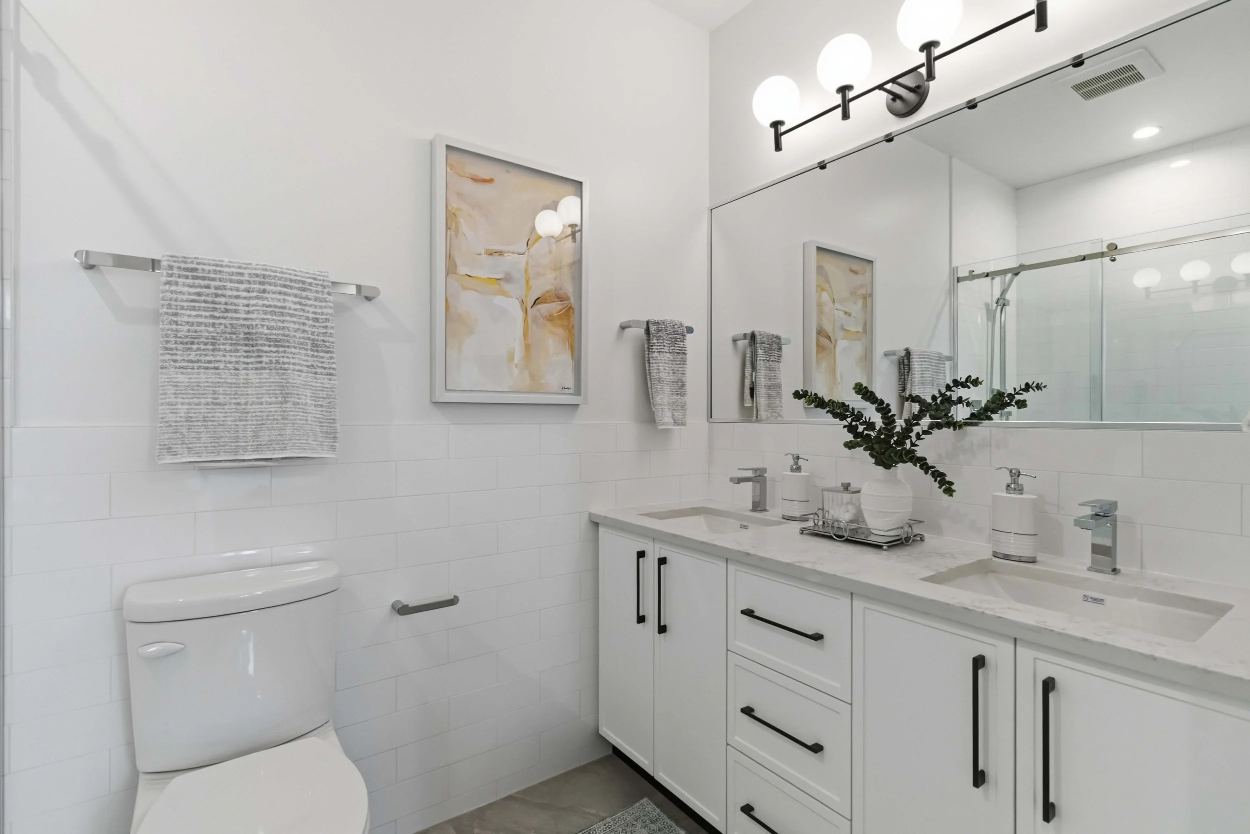 A modern bathroom featuring a white vanity with black handles, a double sink, a large mirror, wall-mounted light fixtures, abstract artwork, towel racks with gray towels, a plant in a white vase, and a glass shower enclosure.