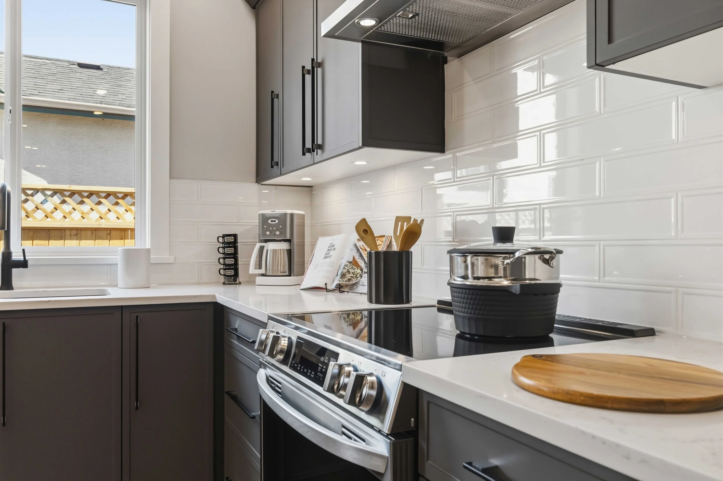 A modern kitchen with gray cabinets, a stove, a white countertop, a window, a coffee maker, a cookbook, utensils in a black holder, and a black pot on the stove.