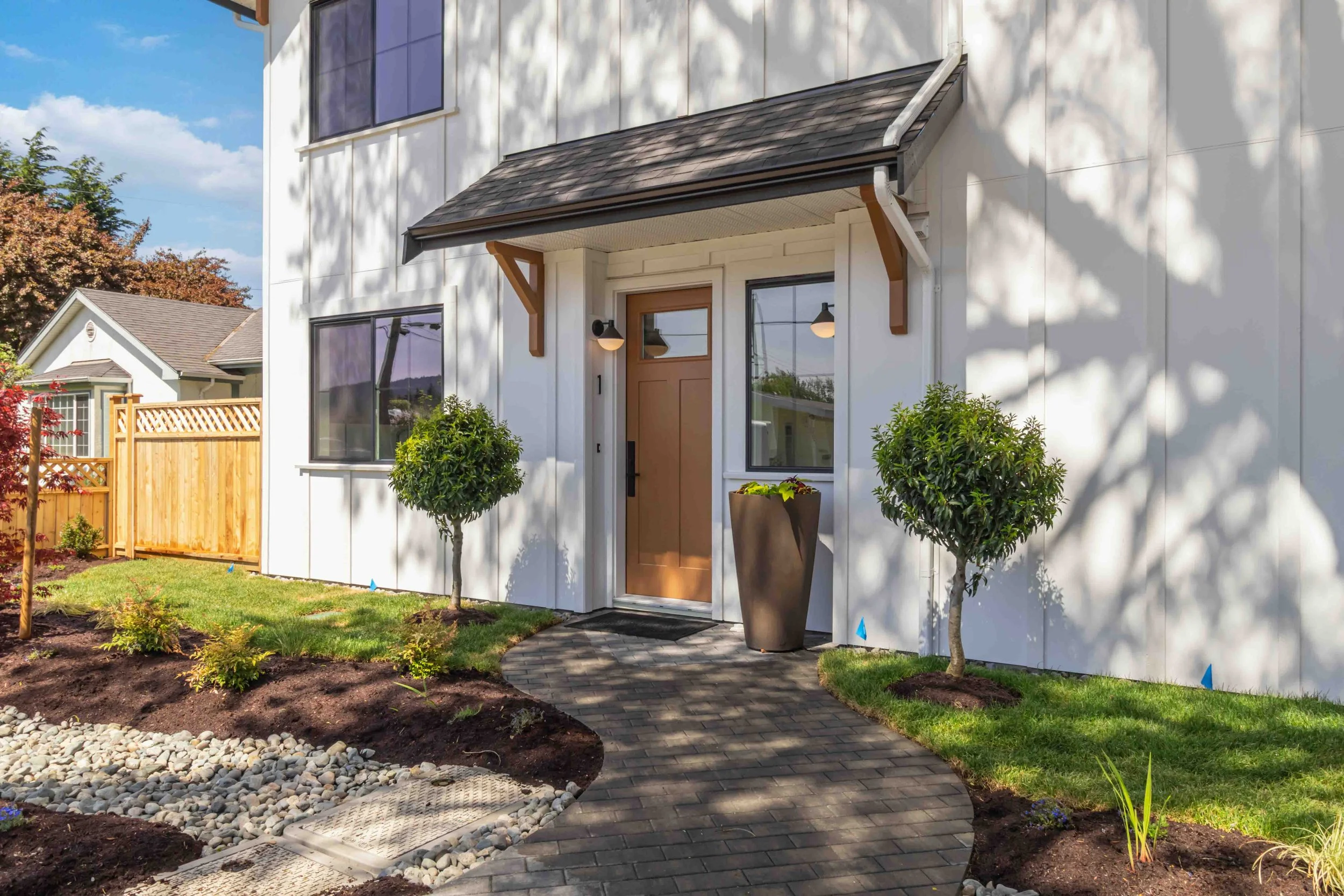 Front entrance of a modern house with a wooden door, potted plants, and a landscaped yard featuring a paved walkway, small trees, bushes, and a garden bed with rocks.
