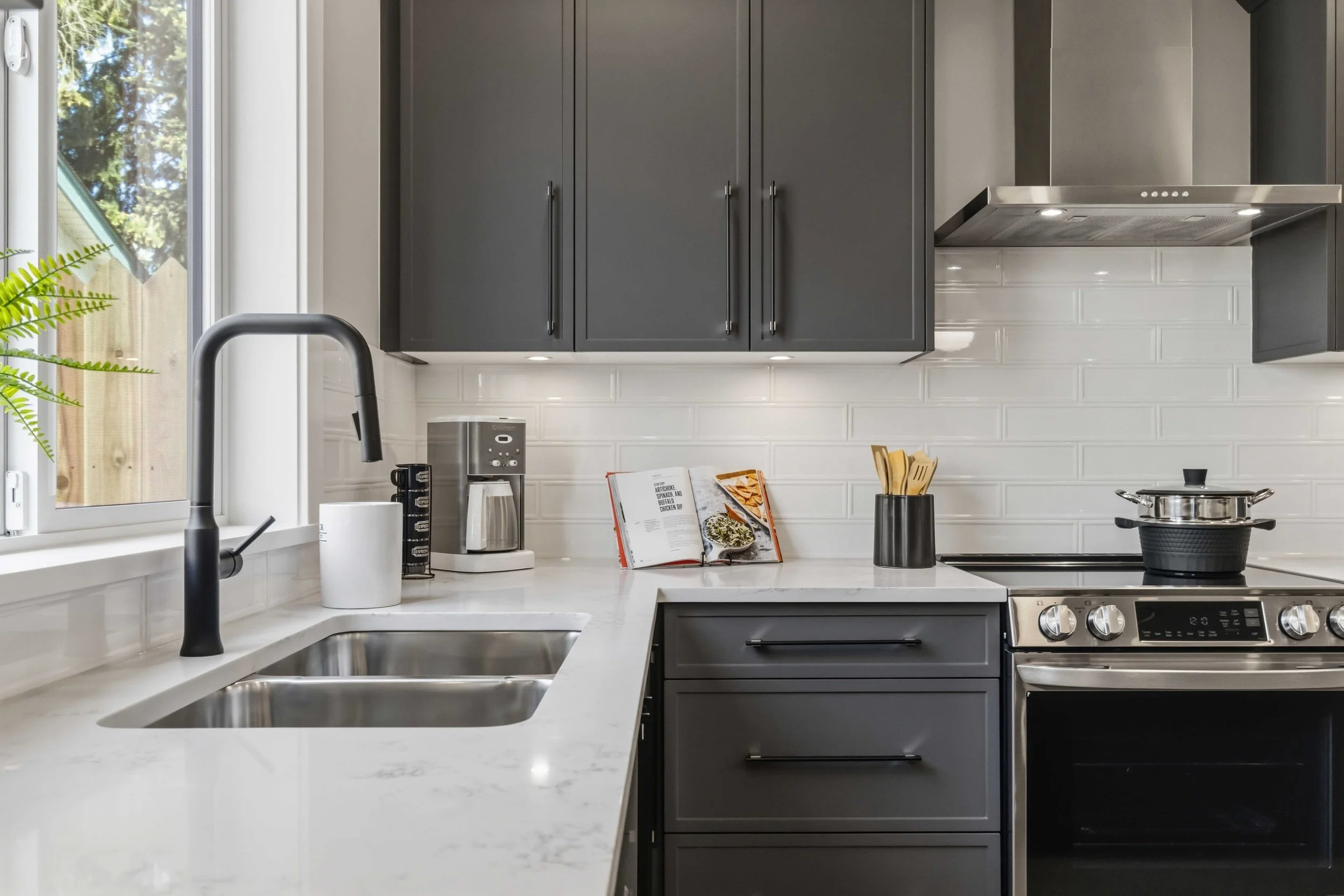 A modern kitchen with gray cabinets, a white marble countertop, and a white subway tile backsplash. There is a window with a view of trees, a black faucet, a coffee maker, an open cookbook, a black utensil holder, and a stove with a pot on it.