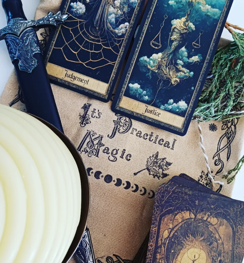A collection of mystical items including a sword with a decorated handle, two tarot cards labeled 'Judgement' and 'Justice' with celestial imagery, a cloth with the phrase 'It's Practical Magic', a green pine branch, and a candle holder with a lit candle.