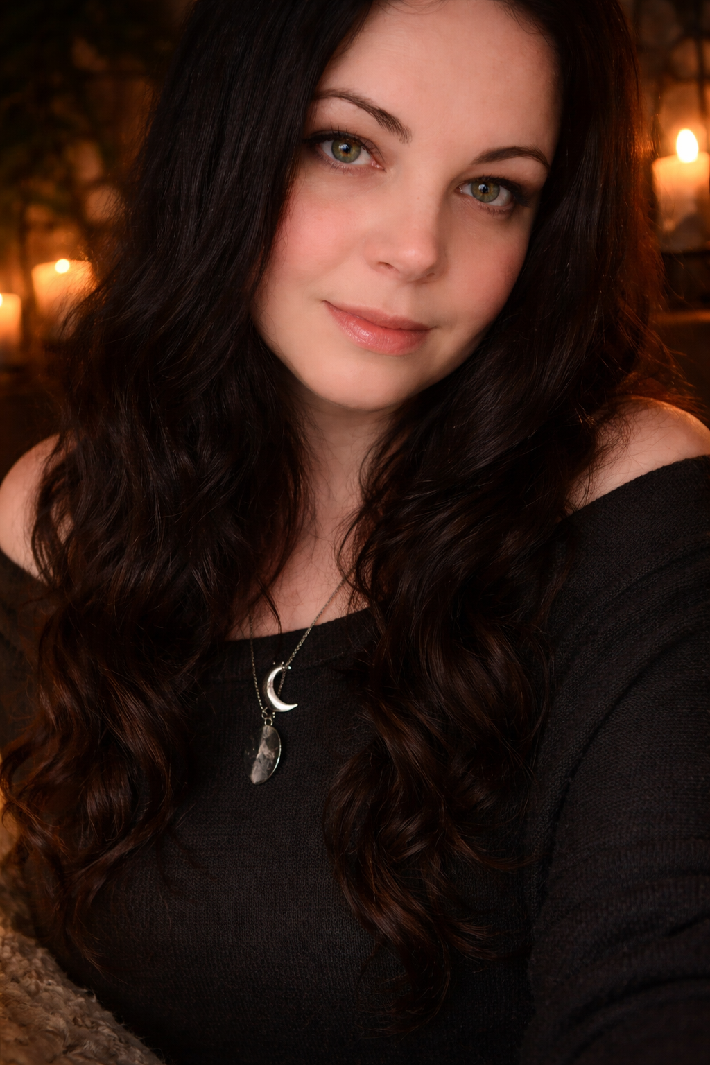 A young woman with long, wavy dark hair and green eyes, wearing a black top and a silver necklace with moon and teardrop-shaped pendants, posing indoors with candles in the background.