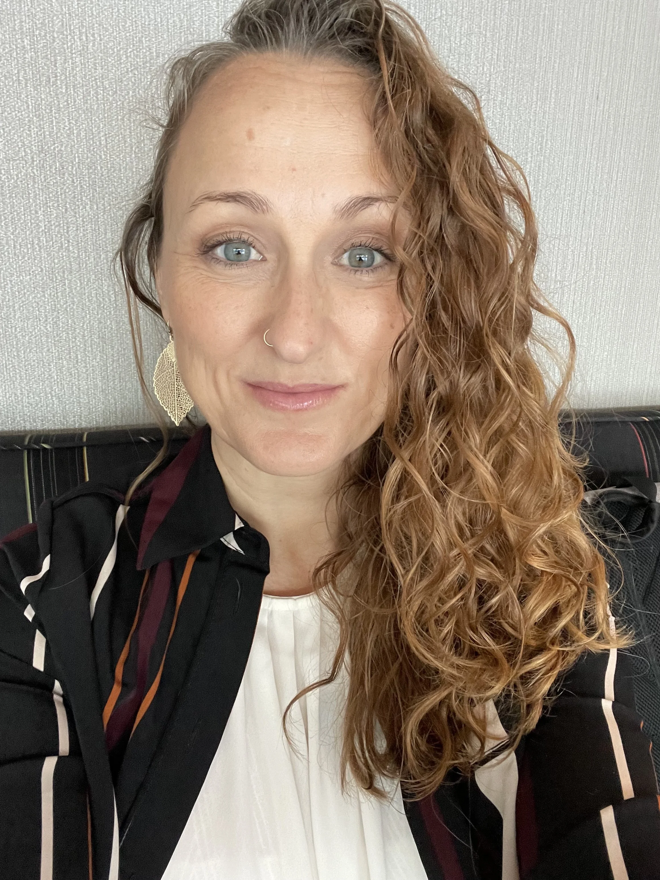 A woman with curly red hair, light blue eyes, wearing large gold earrings and a black and white striped blazer, seated against a plain textured wall.