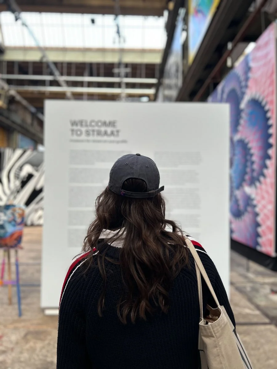 A person with long wavy hair wearing a black cap and a black sweater with red and white stripes on the shoulders, carrying a beige tote bag, stands in front of a large white informational sign in an art gallery or industrial-style venue with artwork 