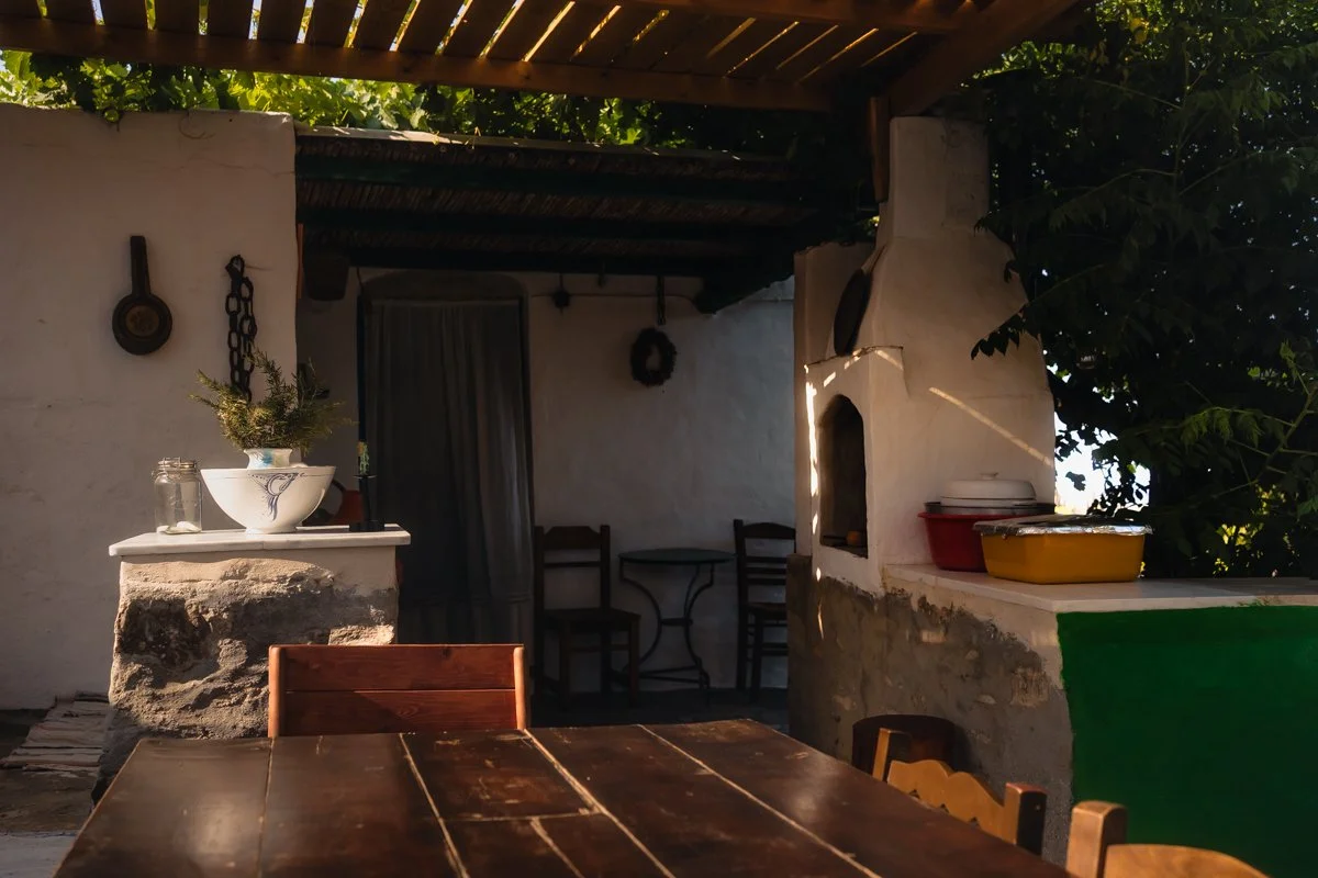 Outdoor patio with a wooden dining table and chairs, a built-in fireplace, hanging decorations, and a small table with a bowl, jars, and containers, surrounded by greenery.