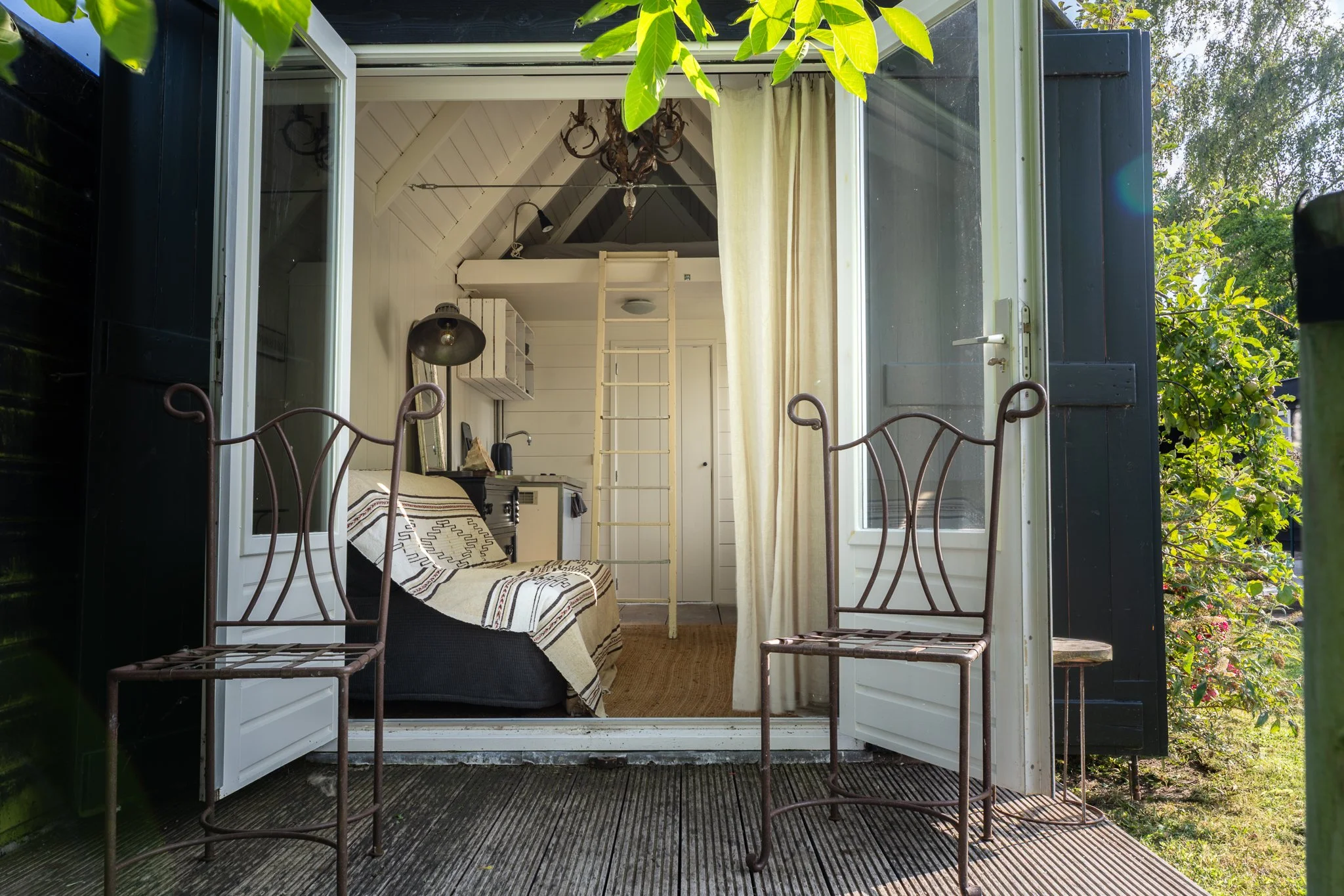 A view of a cozy room with white walls and ceiling, seen through open double doors from a patio with metal chairs. The room features a loft bed with a ladder, a chandelier hanging from the ceiling, and a small workspace with a lamp, a computer, and a