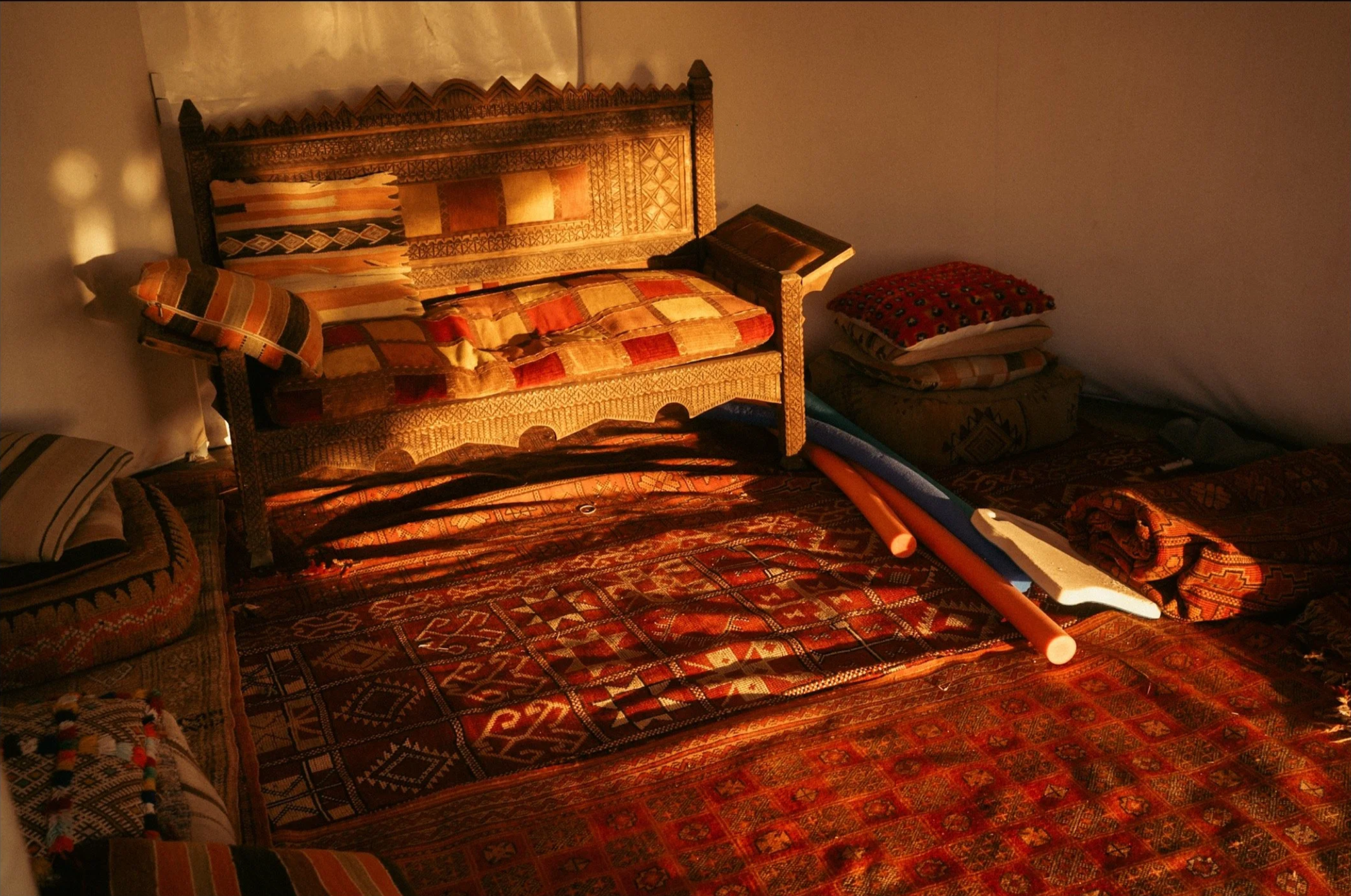 A room with traditional patterned textiles, including a wooden framed sofa, cushions, and a stack of folded blankets on the floor, illuminated by warm light.