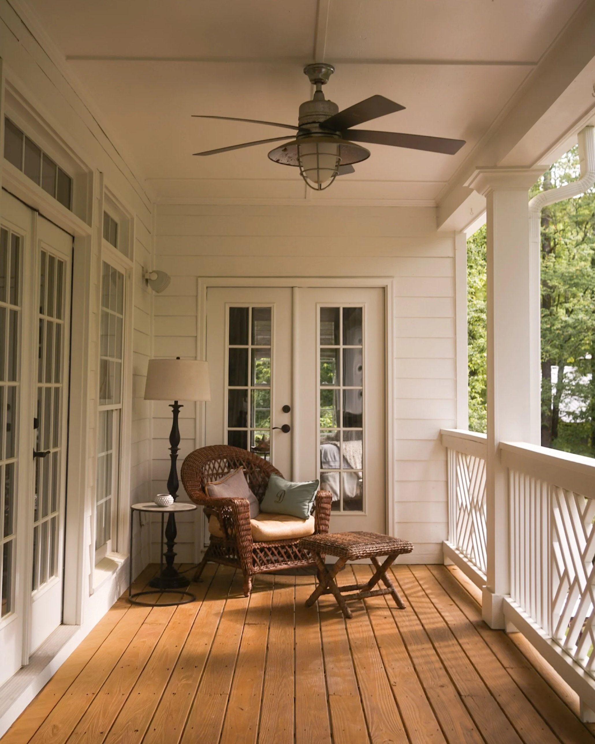 A cozy porch with wooden flooring, white walls, a wicker chair with pillows, a small side table with a decorative item, and a ceiling fan, surrounded by greenery outside.