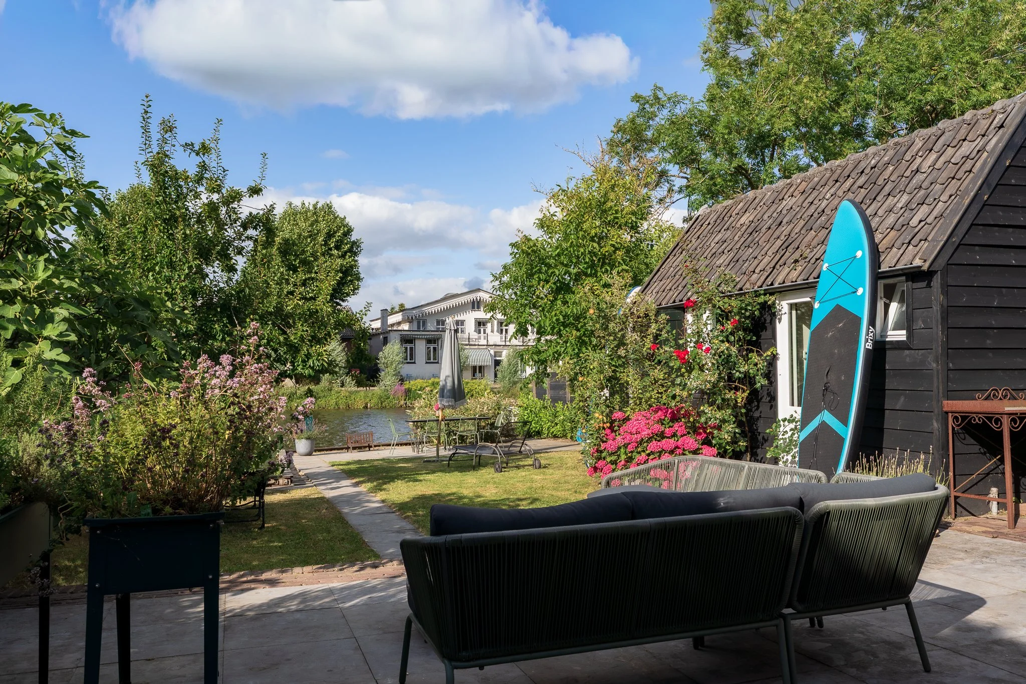 A backyard garden with lush green trees, flowering plants, a pond, and outdoor seating. There's a black wooden shed with a blue surfboard leaning against it, and a white house with a balcony in the background.