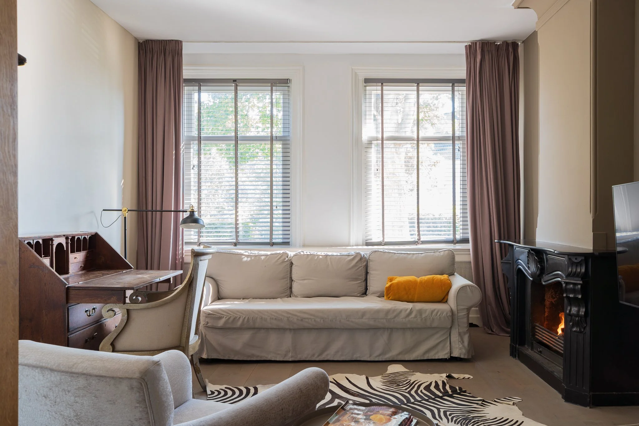 Living room with large windows, beige sofa with a yellow pillow, vintage-style armchair, Zebra rug, fireplace with fire, and a desk with a lamp.