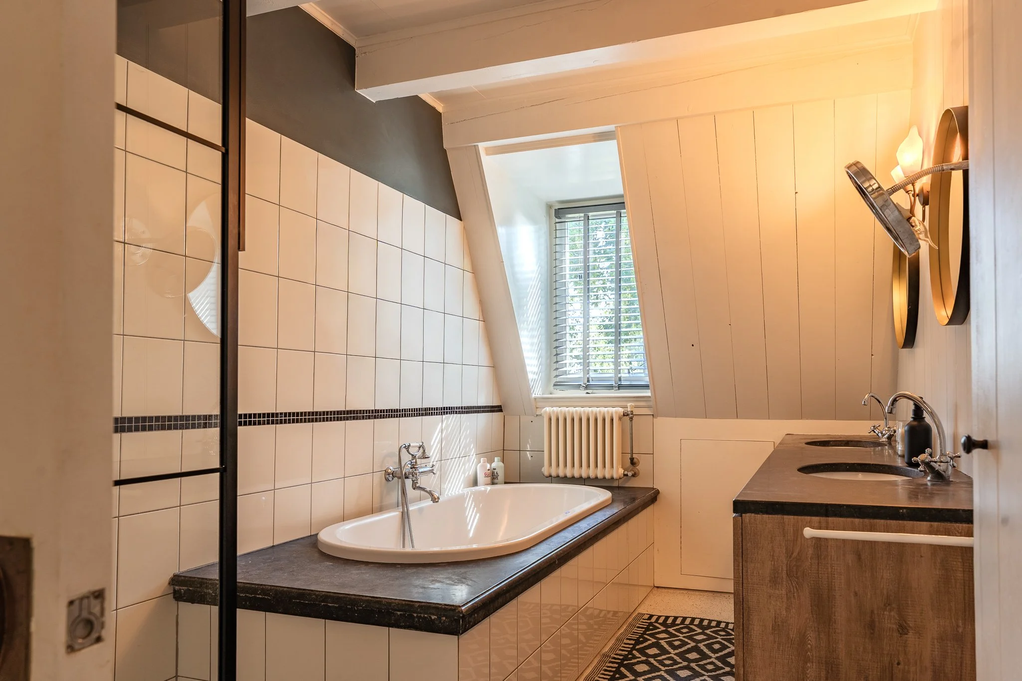 A bathroom with a bathtub, a double sink vanity, a window with blinds, a radiator, and various toiletries.