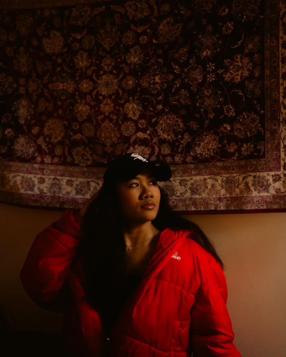 A young woman with dark hair and a black cap, wearing a red jacket, standing in front of a large decorative tapestry with floral patterns.