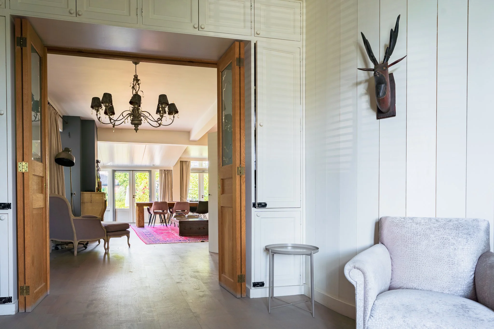 A room with white paneled walls and a beige armchair, visible on the bottom right. An open doorway with wooden trim leads to another room with a chandelier, a pink rug, chairs, and large windows with beige curtains letting in natural light.
