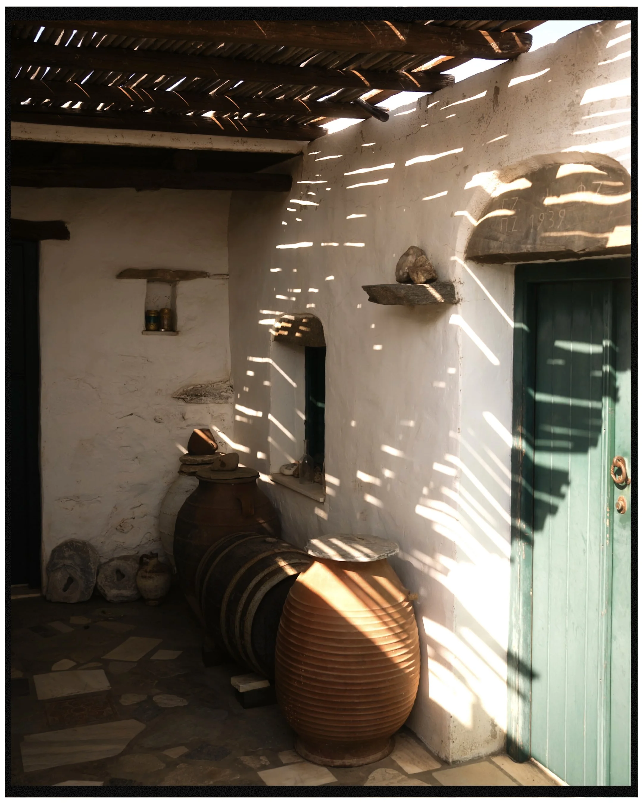A corner of a rustic room with a white plaster wall and a small window. There are large clay pots and several woven baskets stacked on the floor. Two wooden shelves hold some small objects. Sunlight creates patterns of light and shadow on the wall and objects.