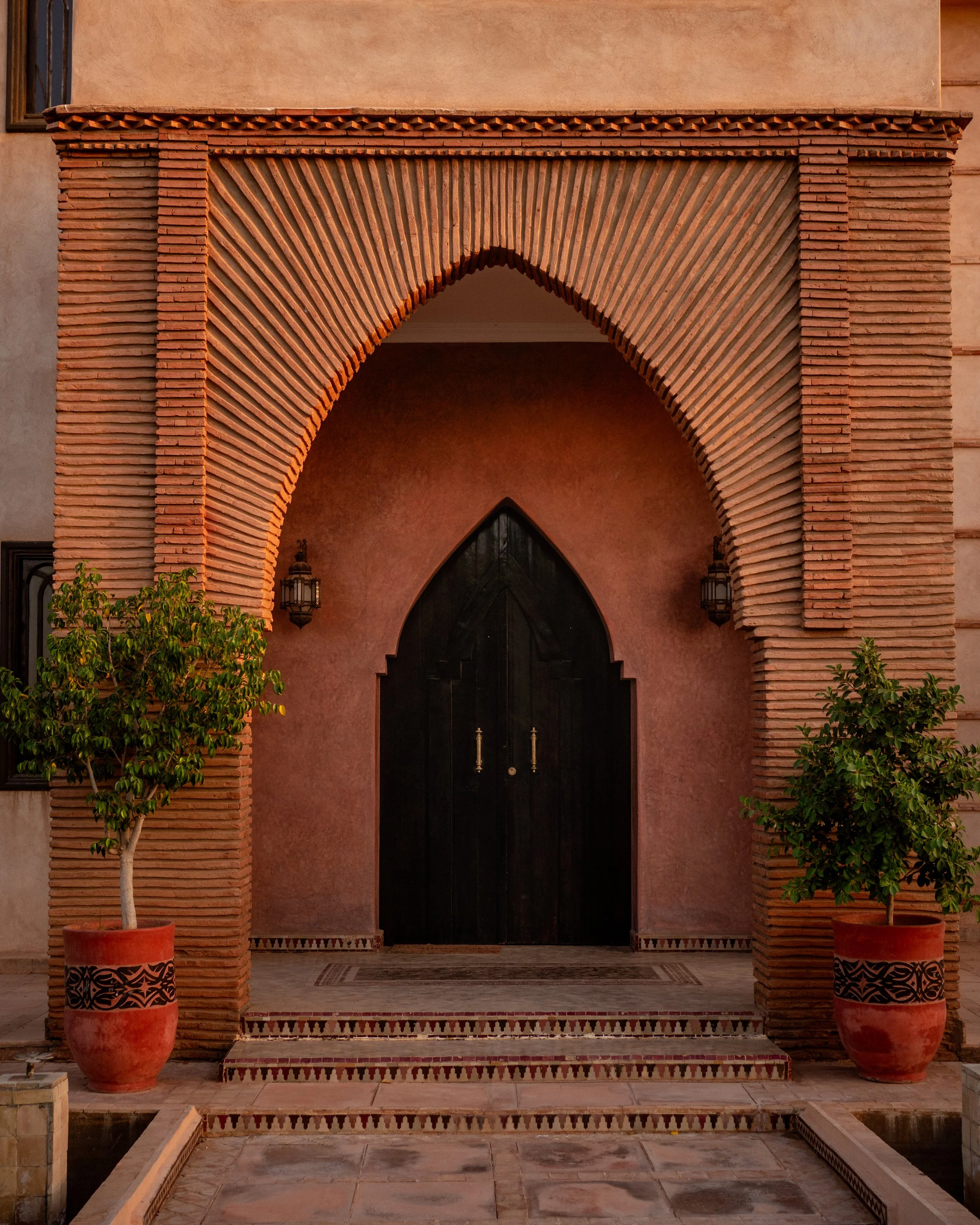 A traditional Moroccan-style archway with brick design, flanked by two potted plants, leading to a black door with lanterns on either side.