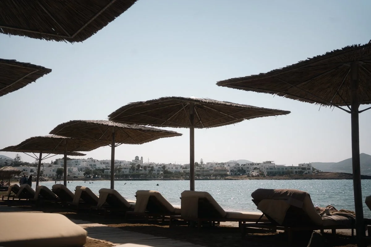 Beach scene with lounge chairs and straw umbrellas overlooking water and white buildings in the distance