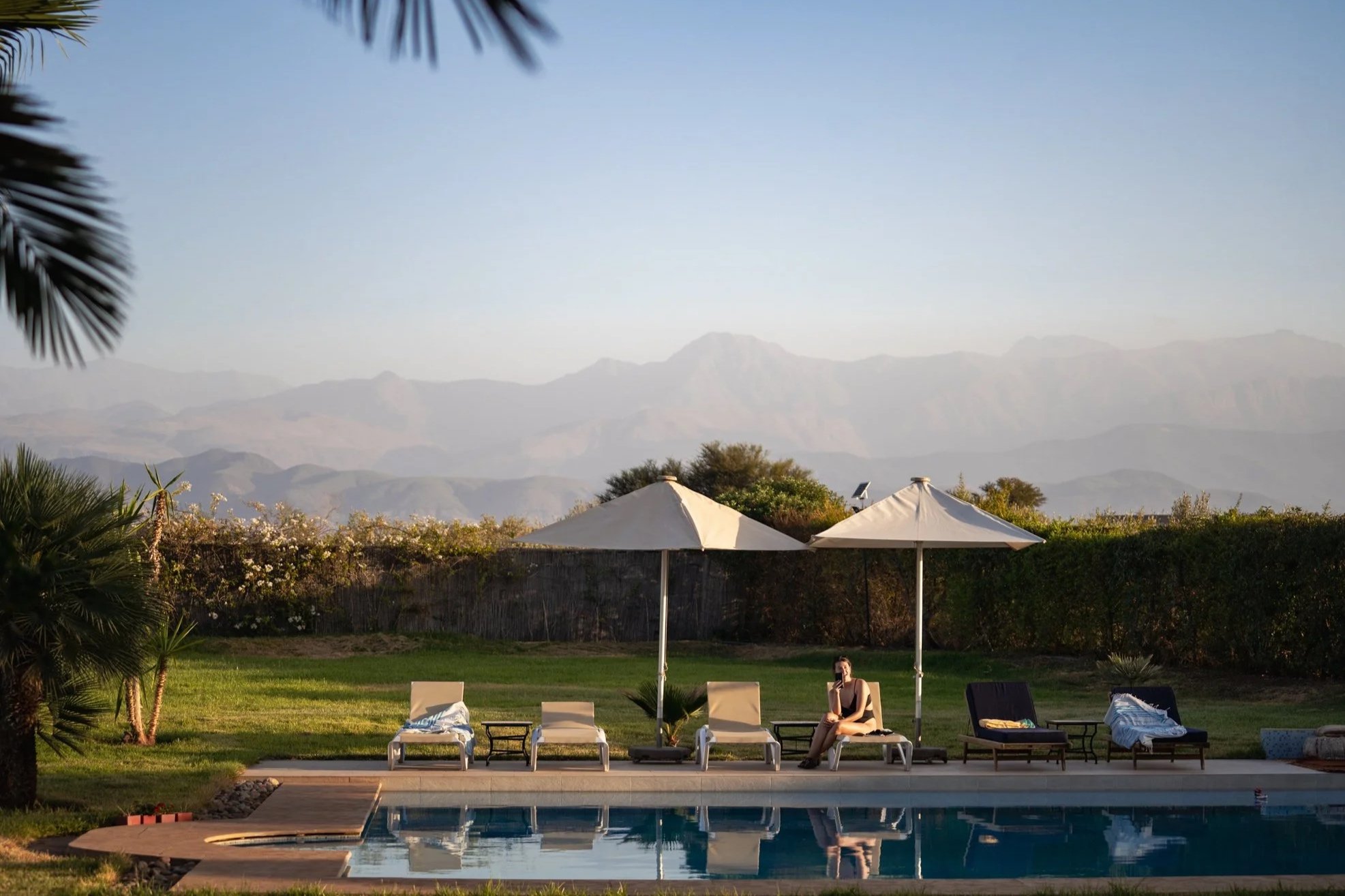 A backyard scene with a swimming pool, poolside chairs, umbrellas, a woman sitting on the chairs, and mountains in the background under a clear sky.