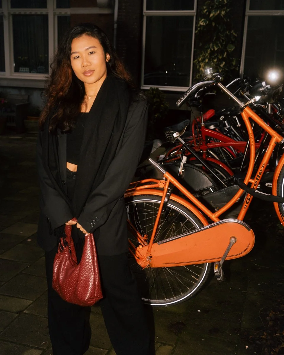 A woman with long dark hair standing outdoors at night, holding a red purse, next to a row of orange bicycles parked on the sidewalk.