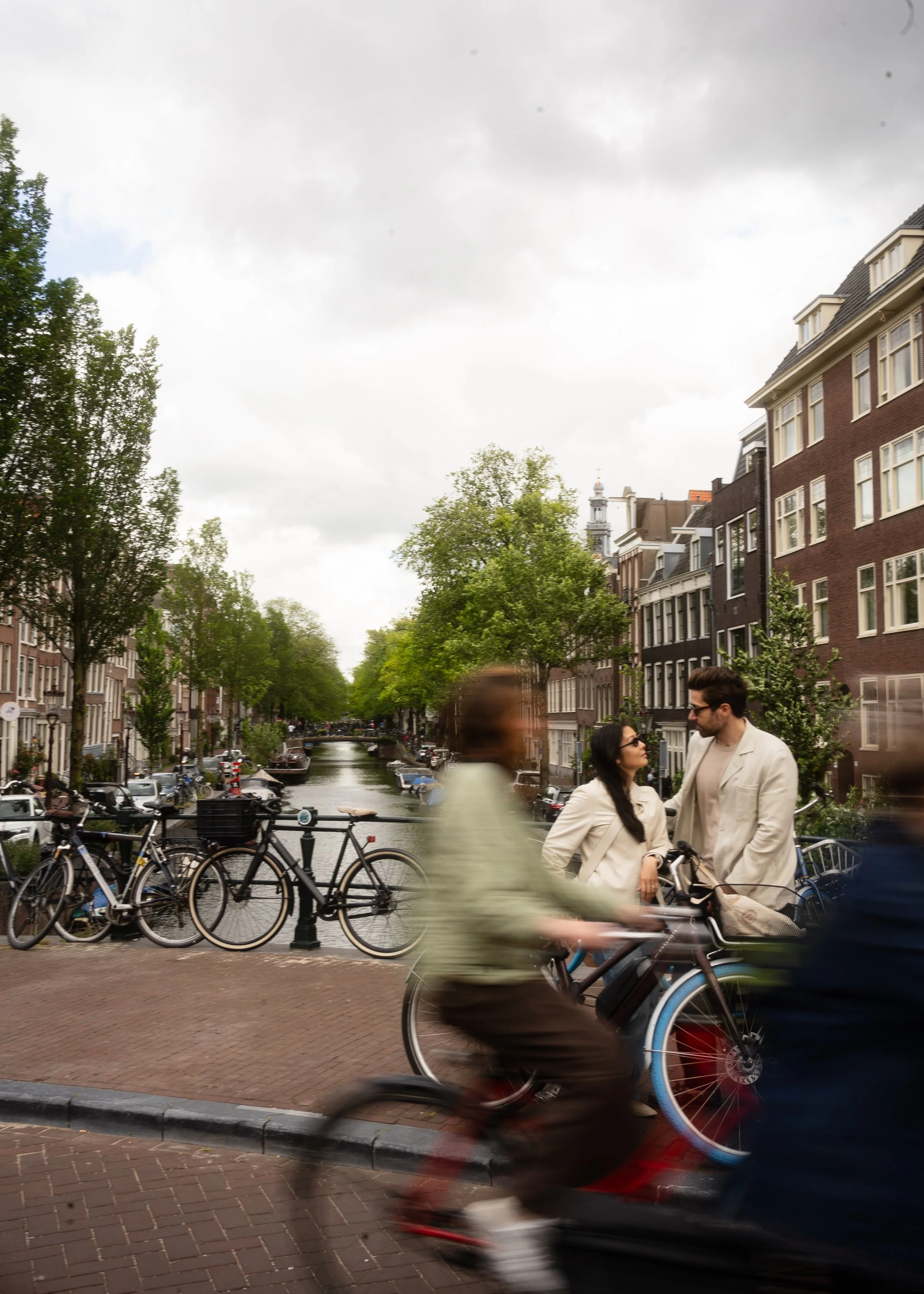 People walking and biking along a canal in a city with traditional European architecture, trees lining the streets, and a cloudy sky.