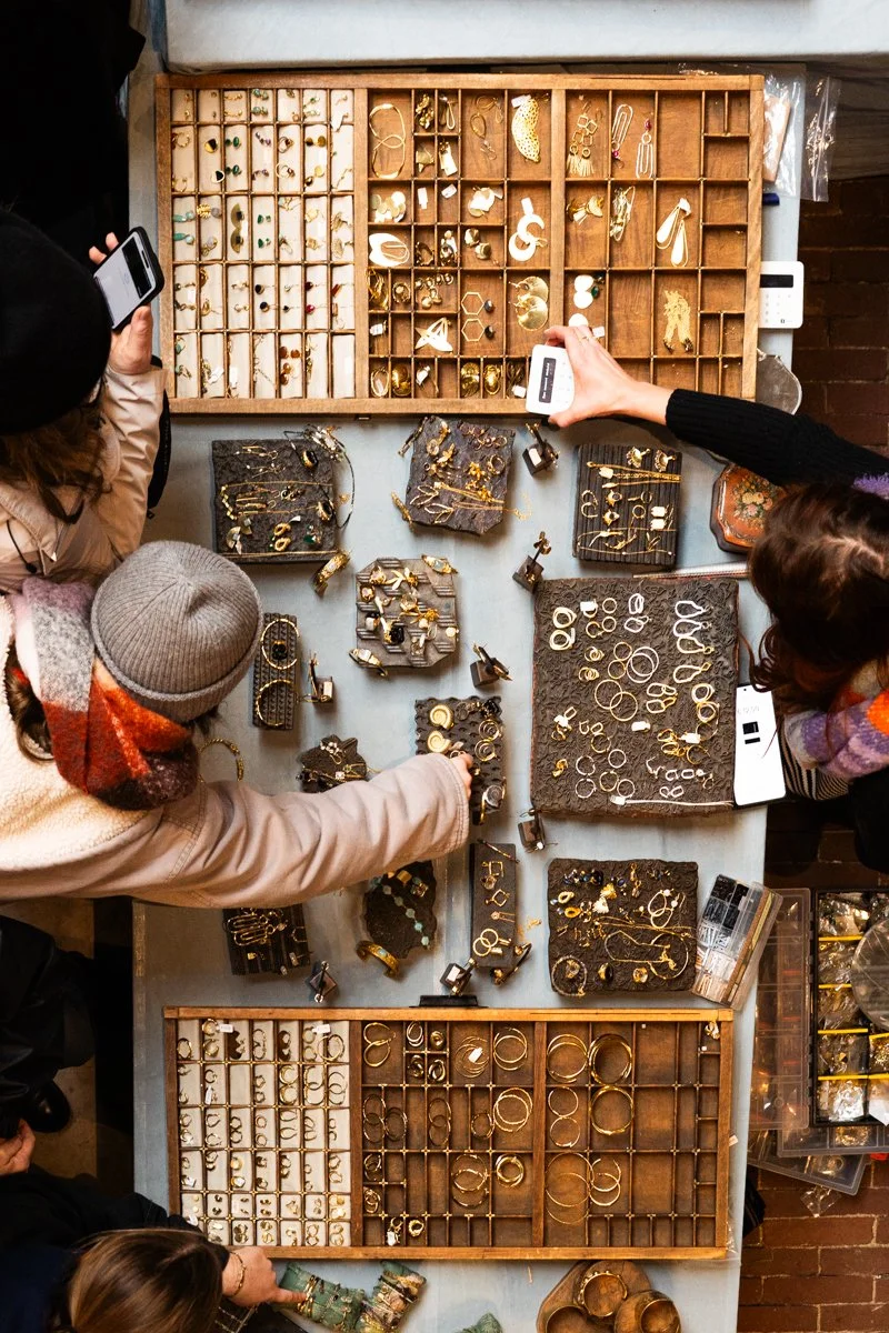 People shopping for gold jewelry at a market stall with assorted rings, earrings, and necklaces displayed in wooden and foam trays.