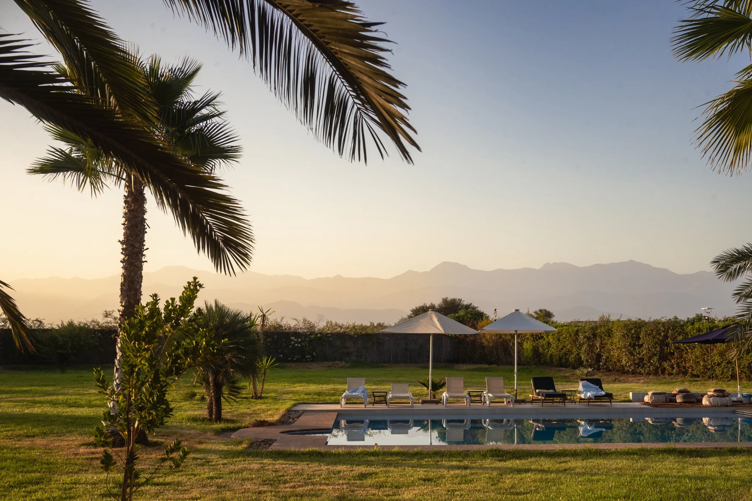 Resort pool area with lounge chairs, umbrellas, and palm trees during sunset, mountains in the background.