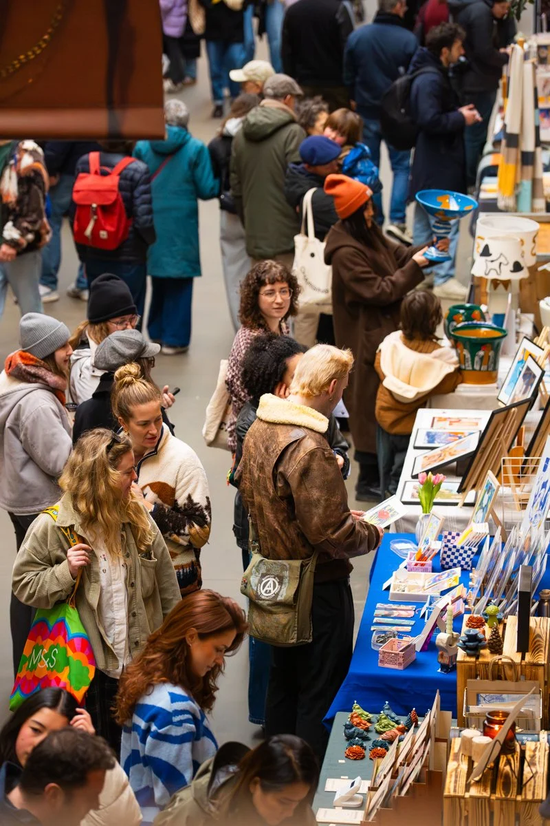 People shopping at an indoor market or craft fair, browsing various art and craft items like paintings and decorative decor.