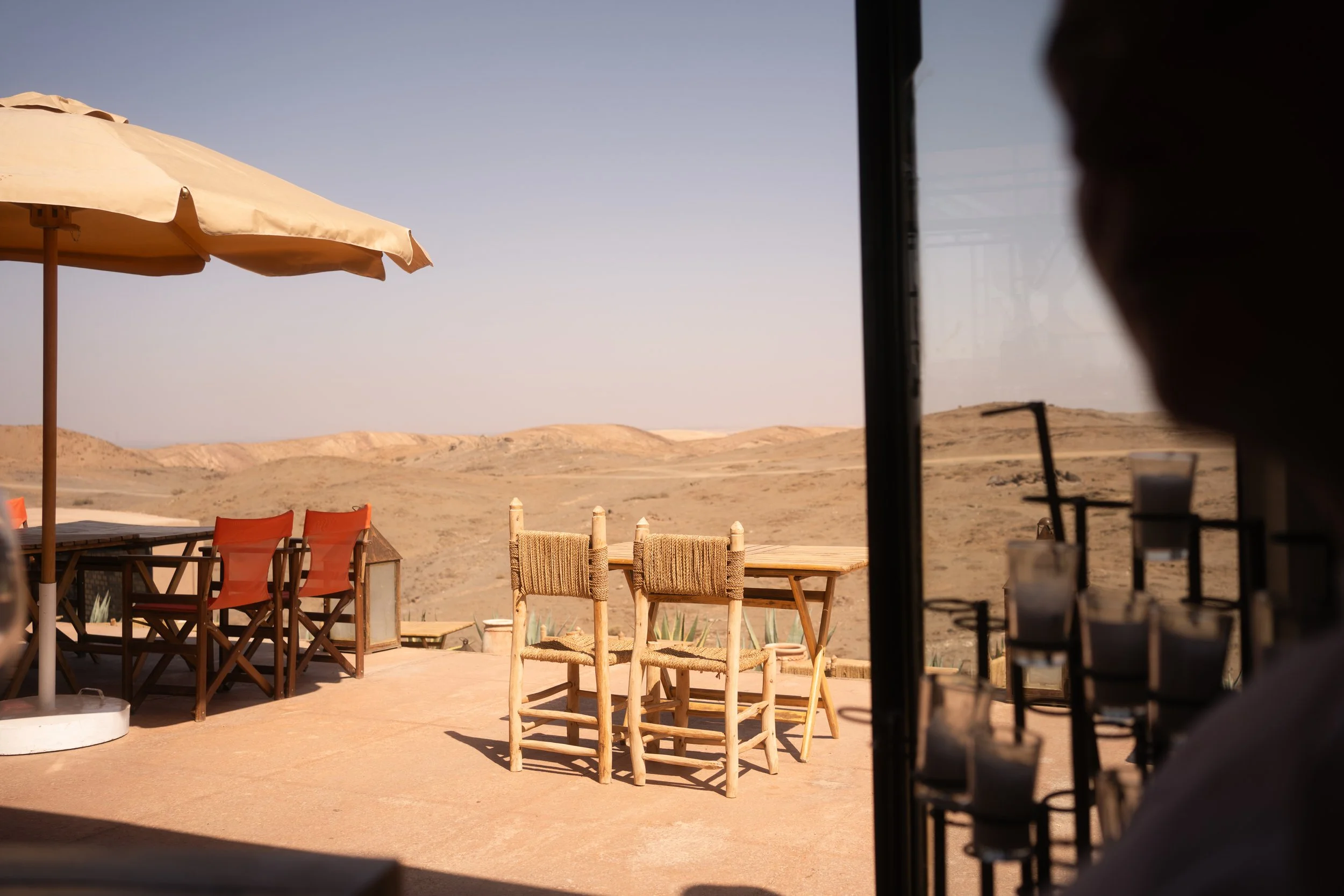 Outdoor seating area with wooden chairs and tables under a large beige umbrella, set against a desert landscape with rolling hills and a clear blue sky.