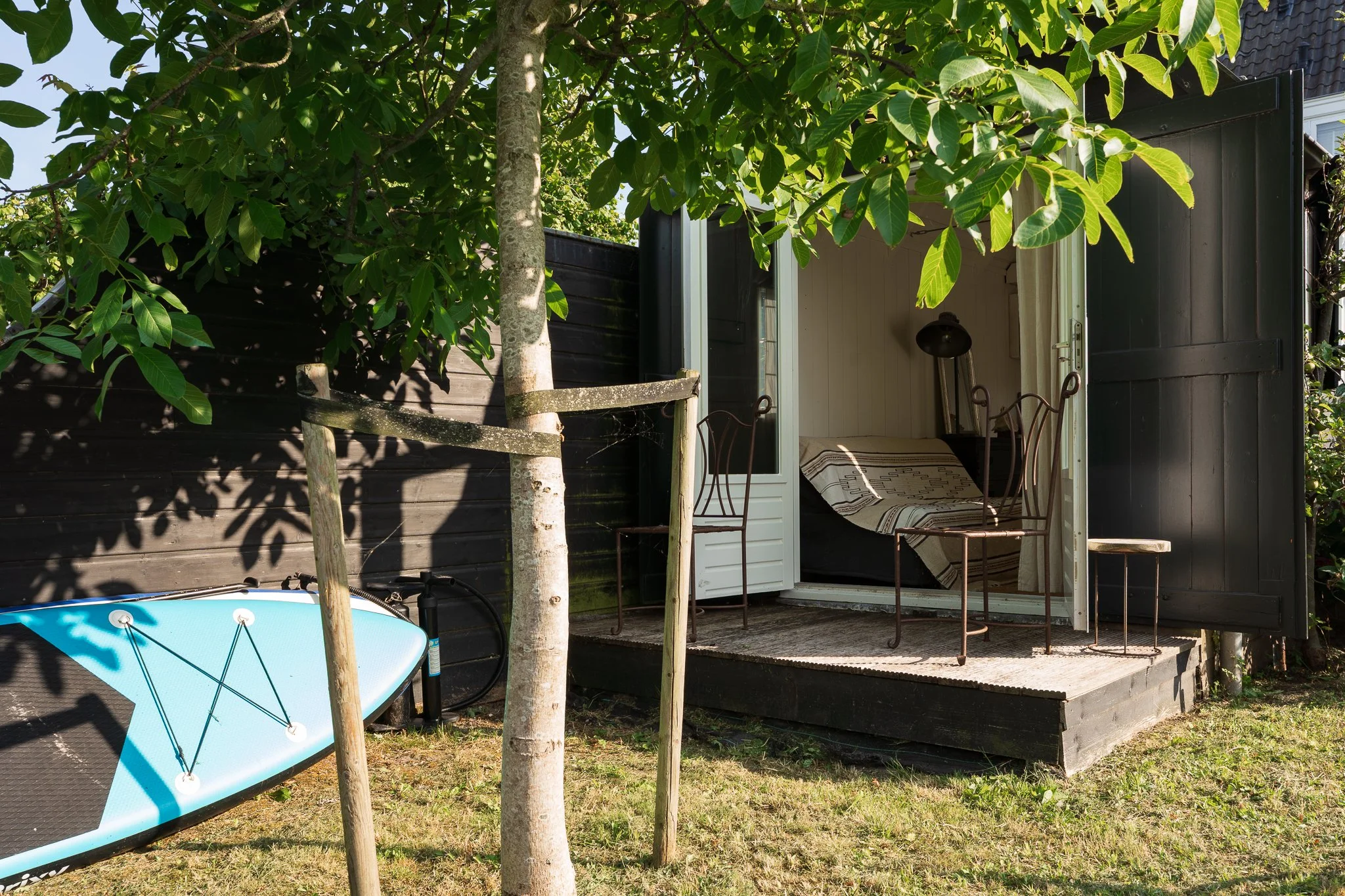 A small outdoor room or shed with an open door, visible interior with a couch and striped blanket, surrounded by green trees and grass, with a blue and black paddleboard and a blue kayak in the yard.
