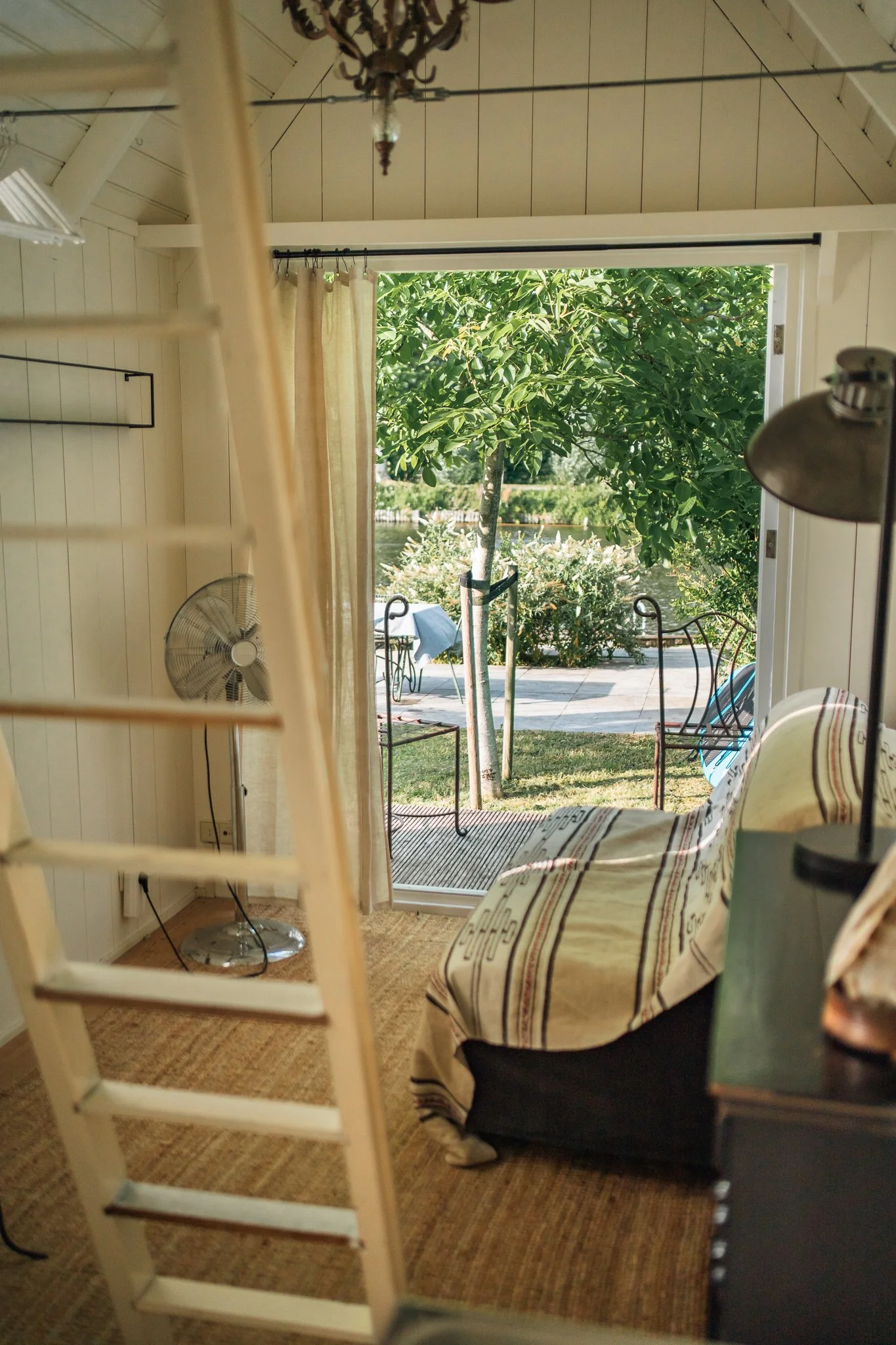 Interior view of a room with a large open door leading to a patio with outdoor chairs and greenery. The room has a lamp, fan, striped fabric on furniture, and a chandelier on the ceiling.