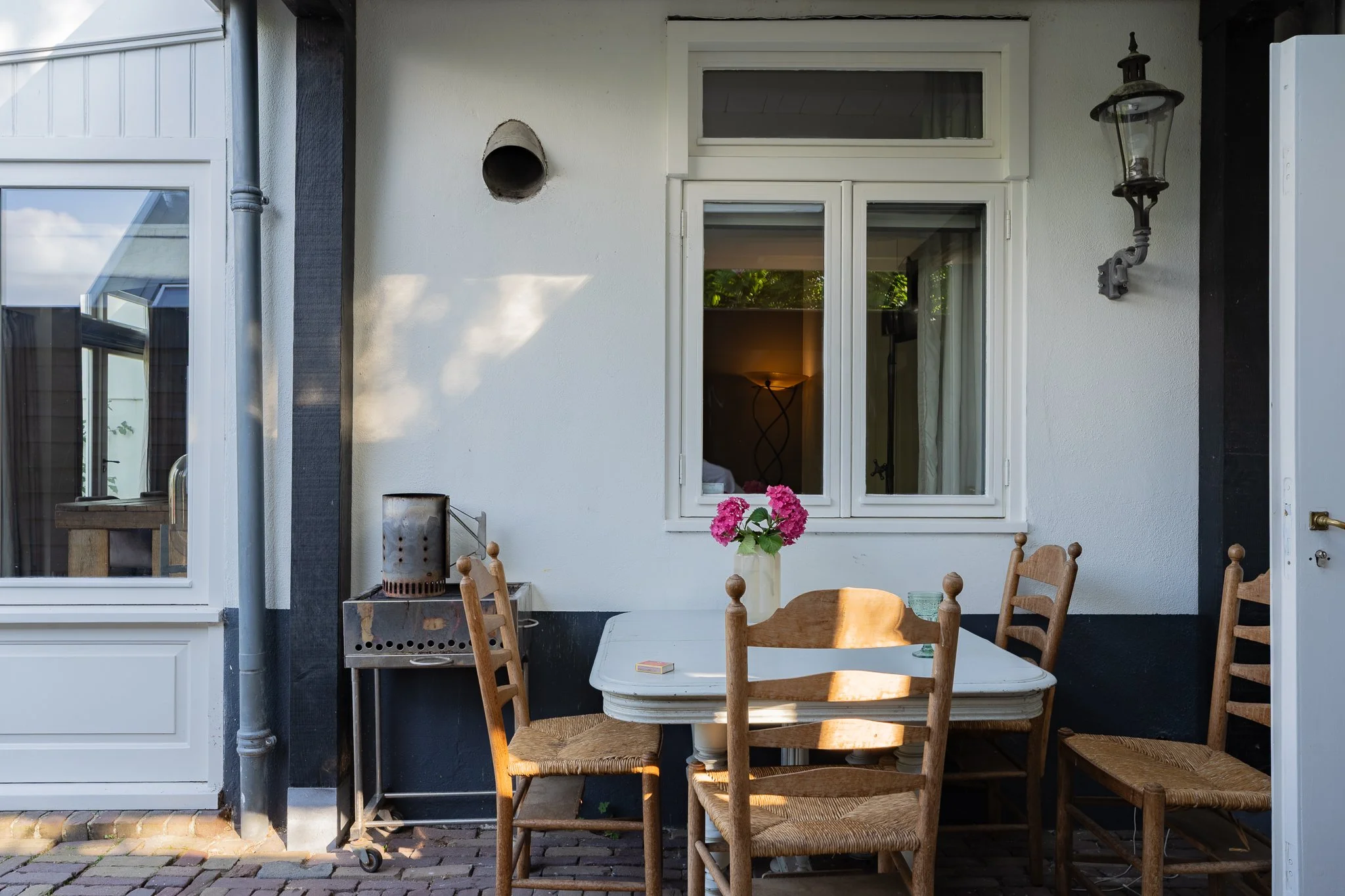 An outdoor patio with a round wooden table and six wooden chairs, a potted pink hydrangea on the table, a vintage lantern, and a small charcoal grill. The white building has a window with a curtain and a ceiling vent, and the patio is paved with bric