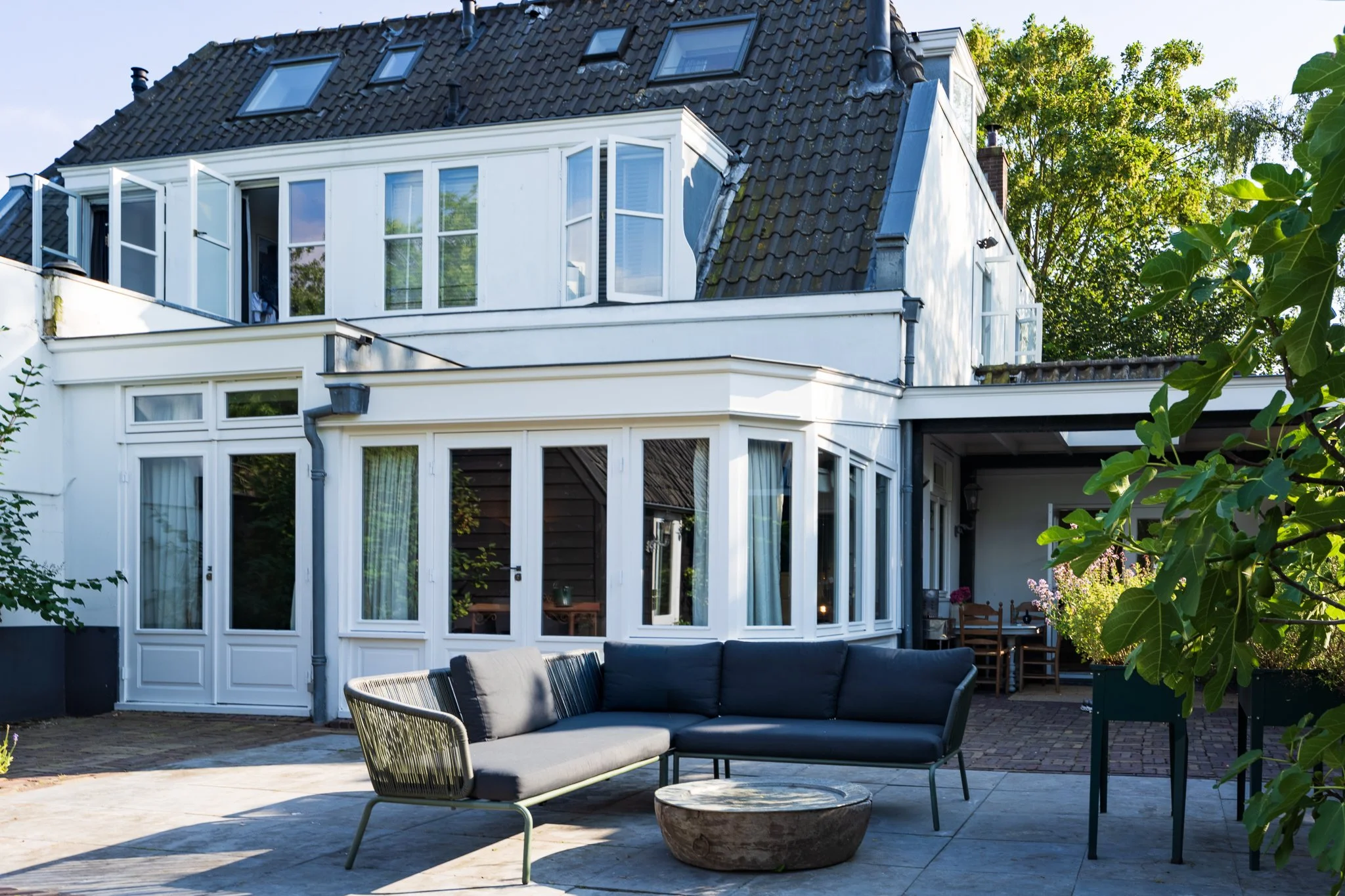 Backyard patio with modern outdoor furniture, including a cushioned lounge chair and small round table, in front of a white house with large windows and a slate-tiled roof with skylights, surrounded by greenery.