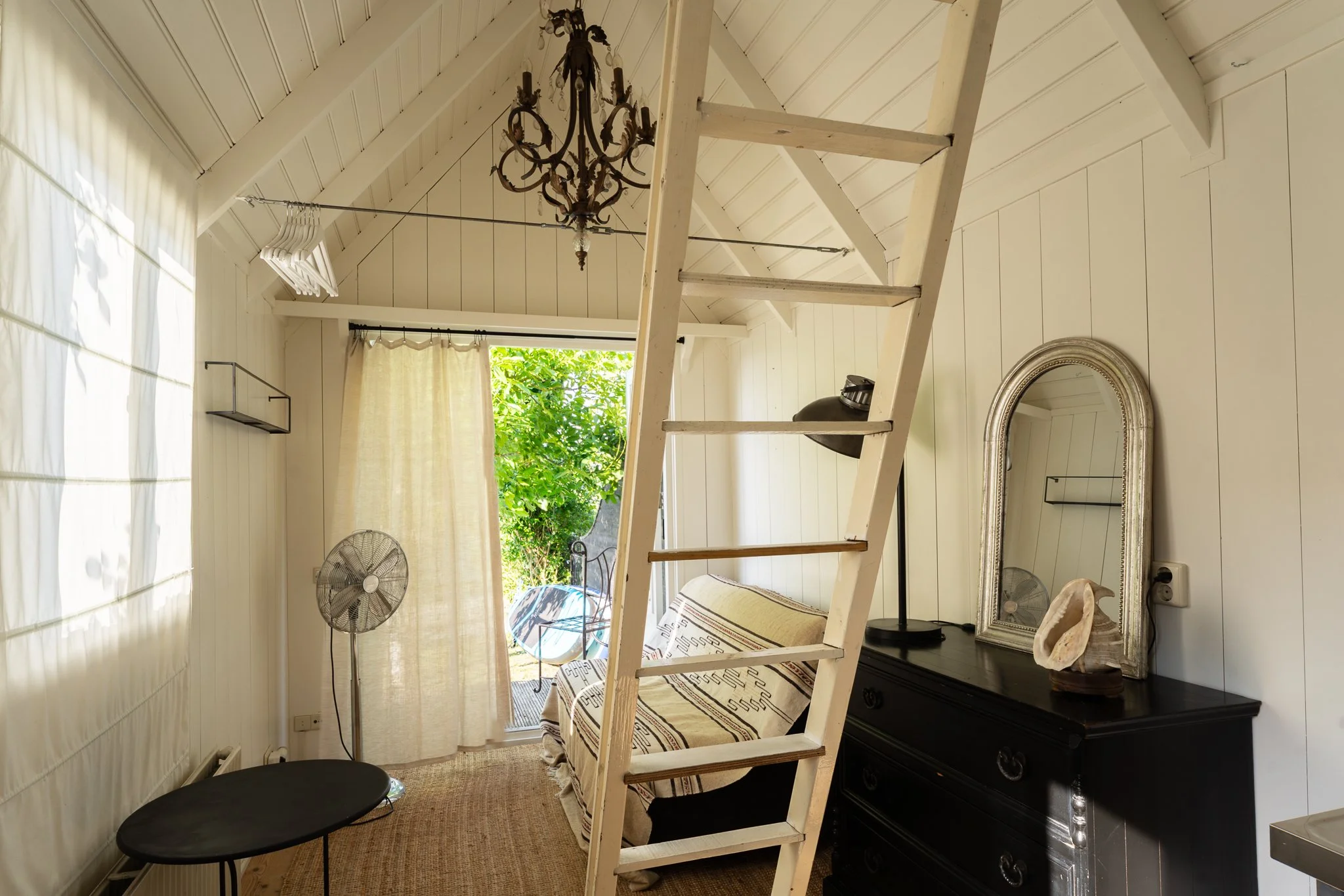 A cozy room with white walls and a slanted ceiling, featuring a ladder, a chandelier, a mirror, a window with open curtains, a black dresser, and decorative items.