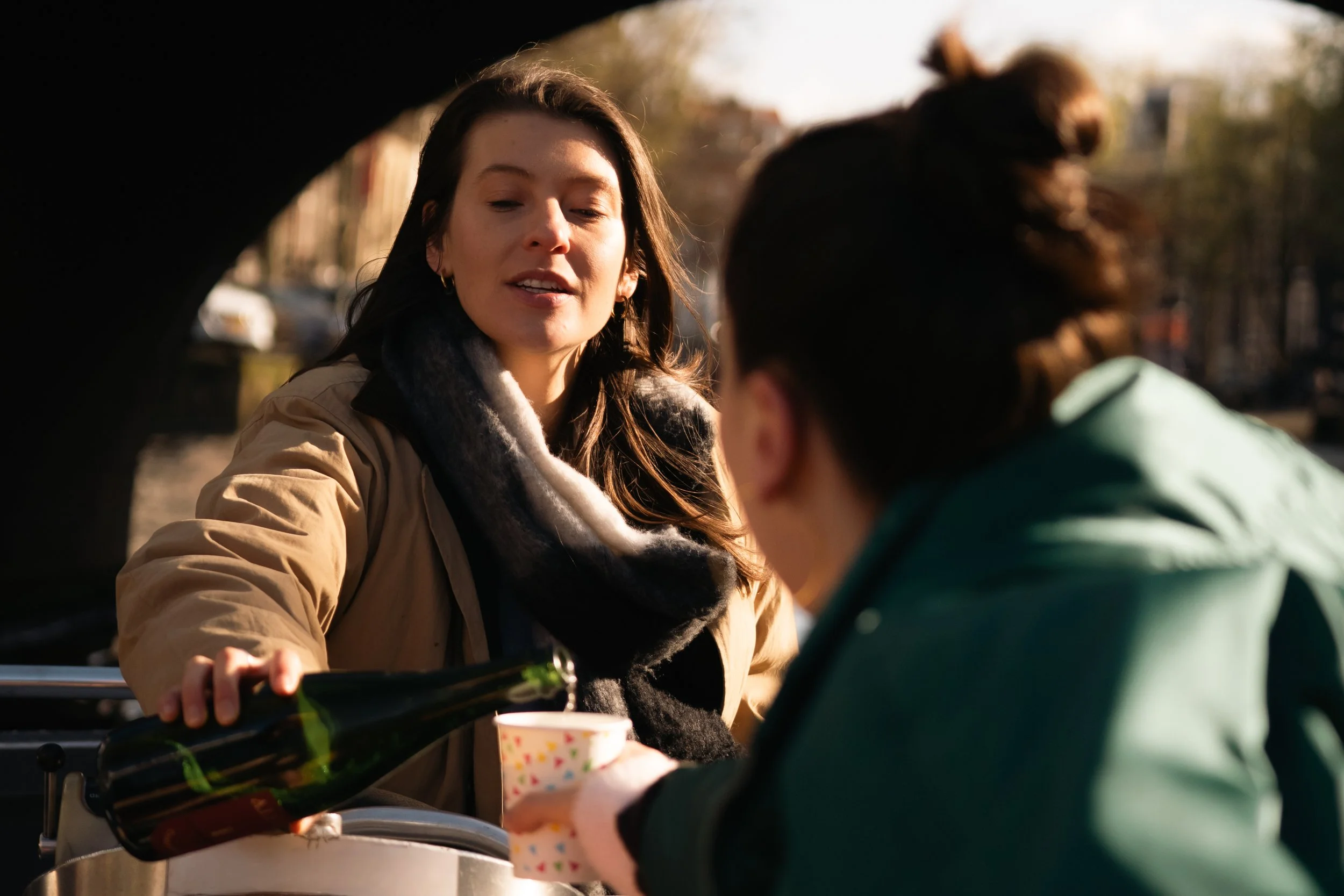 A woman with long dark hair and wearing a beige jacket and black scarf, serving drinks to a person in a green jacket, outdoors on a sunny day.