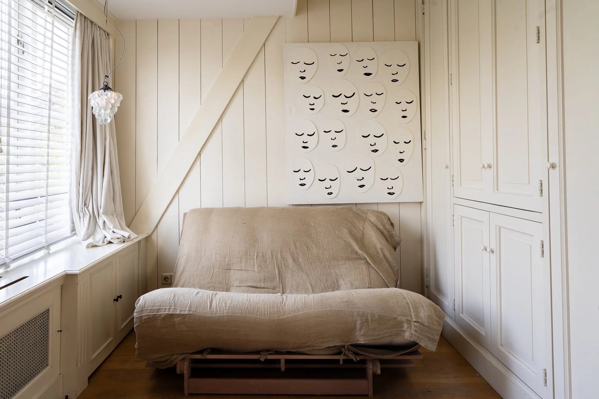 A bedroom with white paneled walls, a window with white blinds and curtains, a beige bed with a mattress without bedding, a white decorative wall art with faces, and built-in white cabinets.