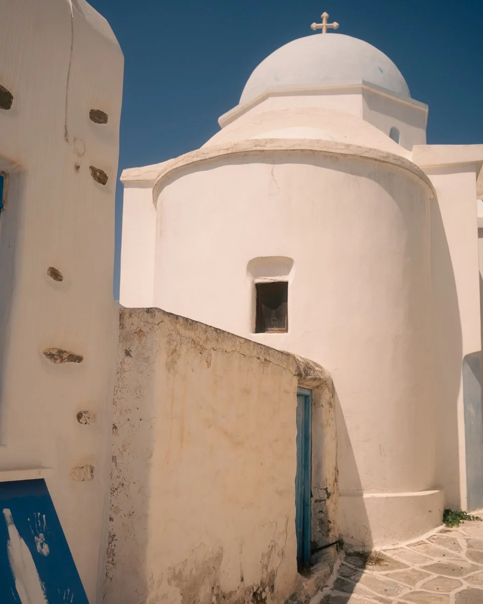 Whitewashed chapel with a dome and cross on top, typical of Greek island architecture, set against a clear blue sky.