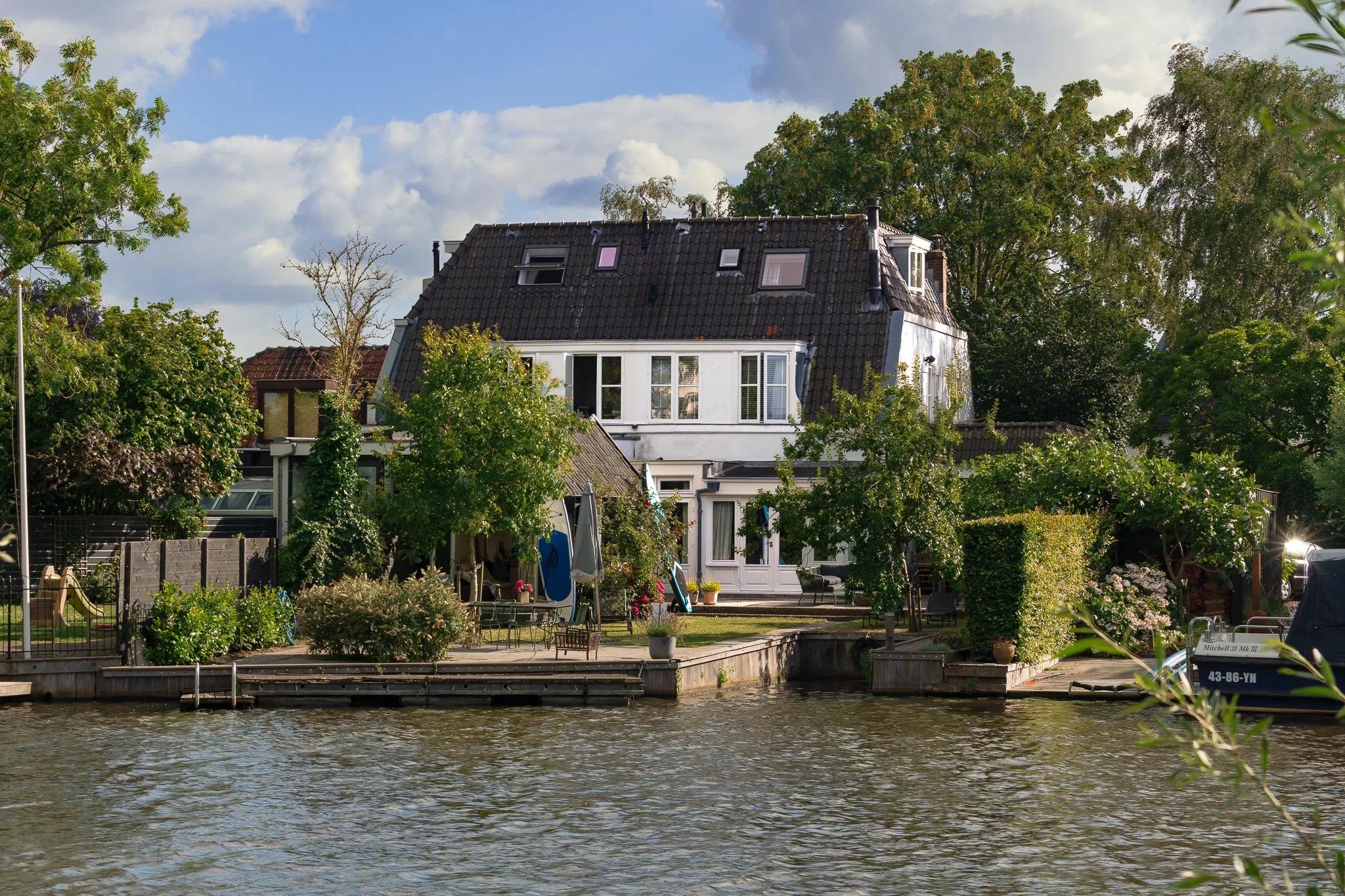 A house with a dark tiled roof and multiple windows, surrounded by trees and greenery along a waterway, under a partly cloudy sky.