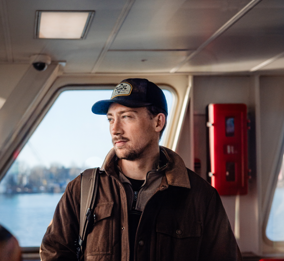 A man with a backpack on a boat, wearing a baseball cap and brown jacket, looking out the window on a sunny day.