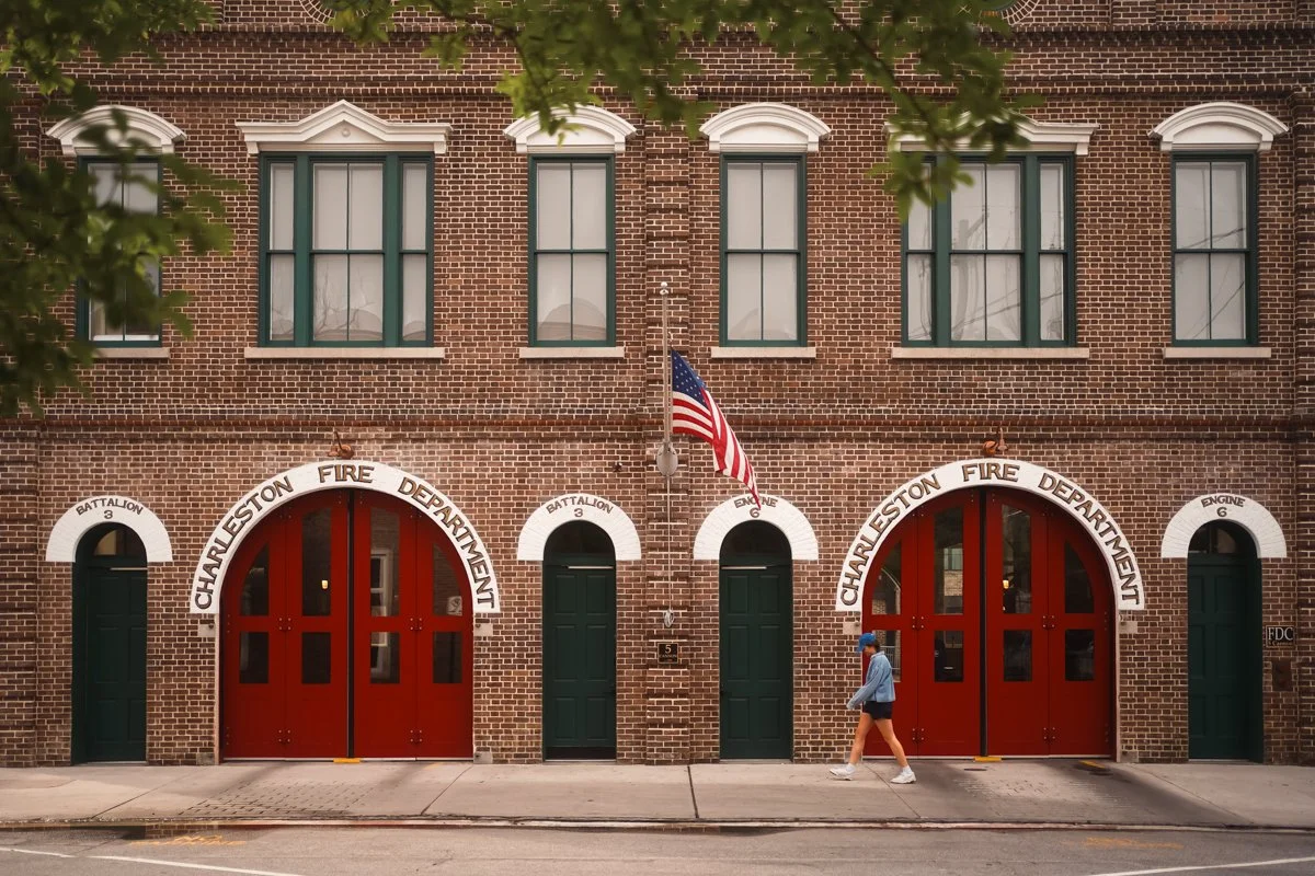 The Charleston Fire Department station building made of red brick with large red arched garage doors, green side doors, and signs indicating Battalion 3 and Engine 6. An American flag is mounted in the center above the garage doors. A person is walki