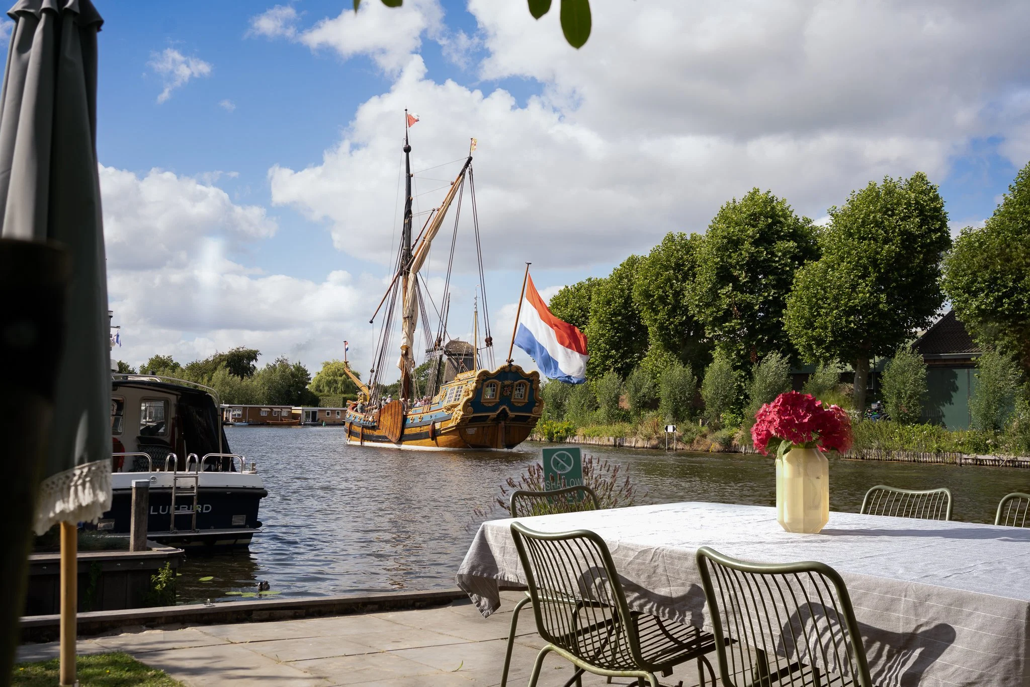 A scenic view of a canal with a decorated wooden sailing ship with flags, water, and lush green trees on the opposite bank. In the foreground, there is a table with a vase of pink flowers and chairs, with a cloudy sky overhead.