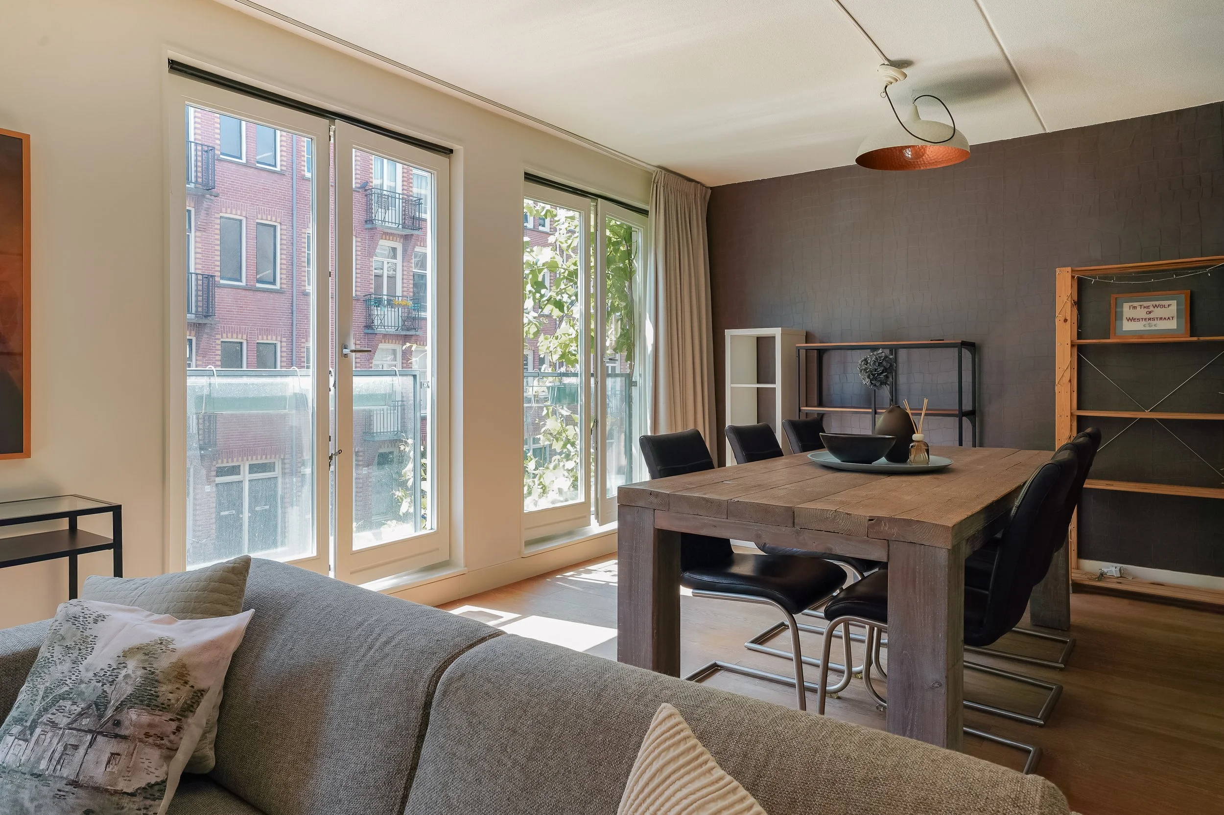 Interior view of a living and dining area with large windows, a wooden dining table with black chairs, a gray couch with patterned pillows, and existing furniture including shelves and a chalkboard, with neighboring building visible through the windo