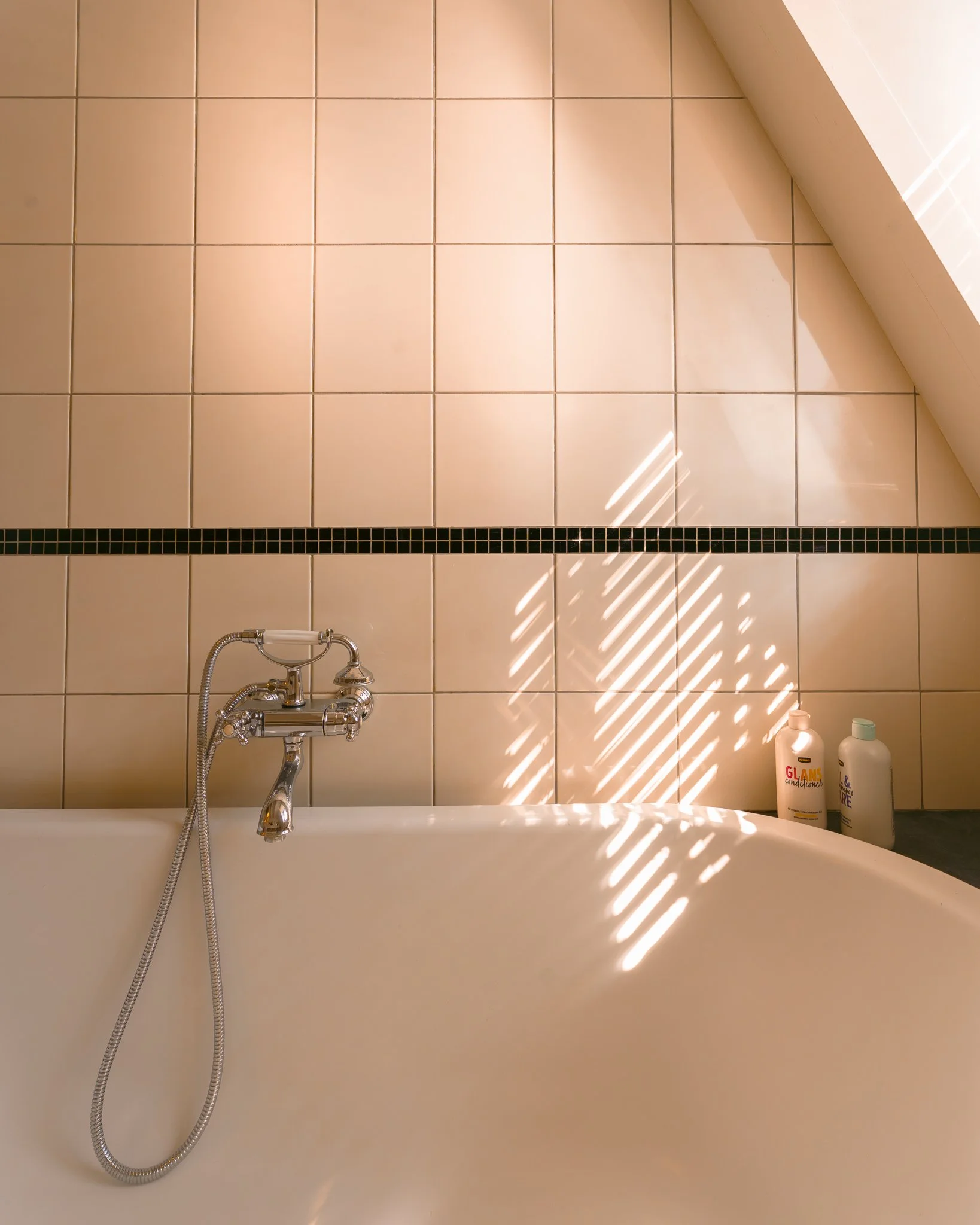 A bathroom with beige tiled walls, a bathtub, a silver faucet and shower hose, and two bottles of shampoo or conditioner on the edge of the tub. Sunlight is casting patterns through a window with blinds onto the wall.