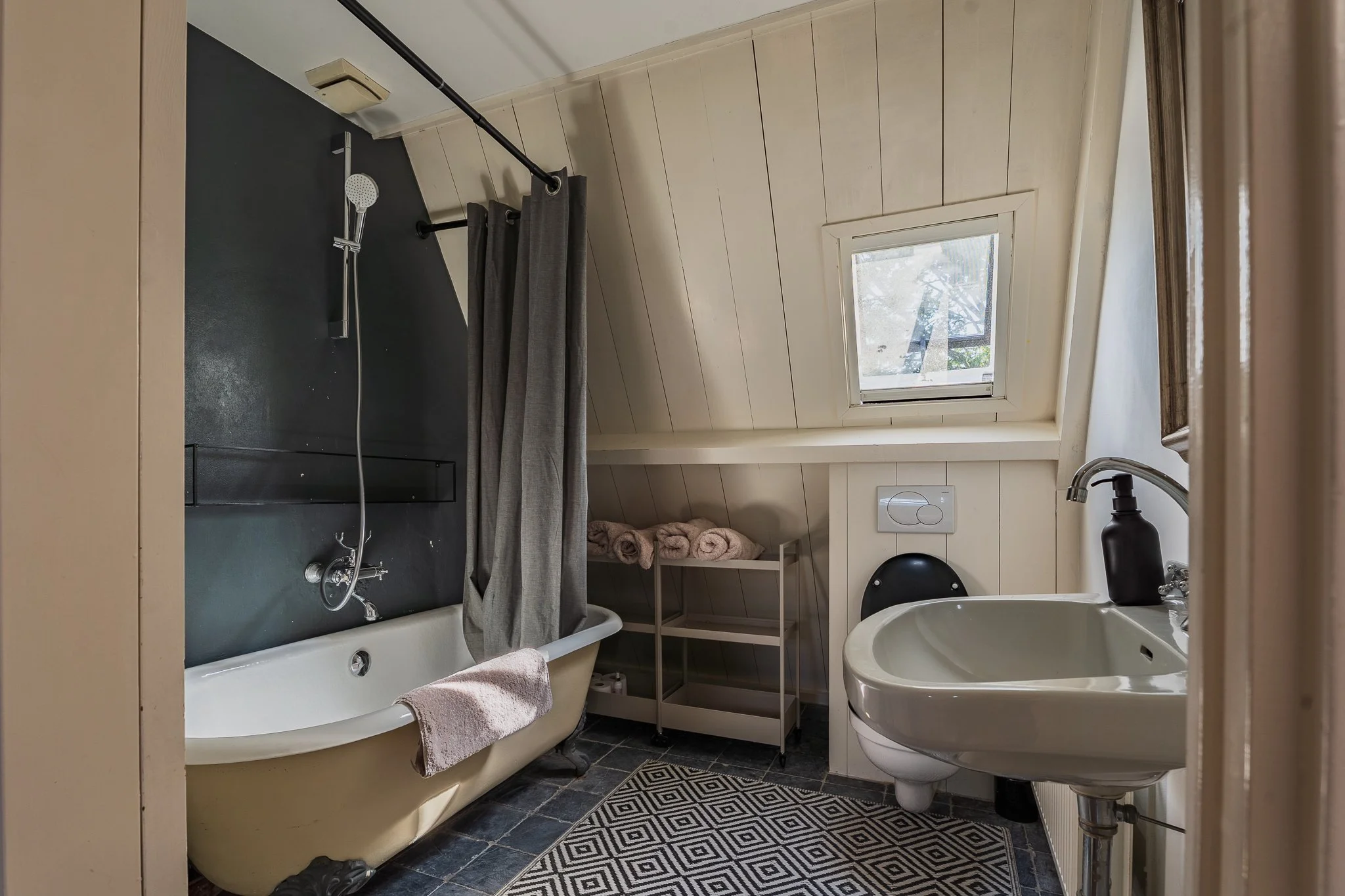 A cozy bathroom with a vintage bathtub and shower, a small window letting in natural light, and a black and white geometric rug on the dark tile floor