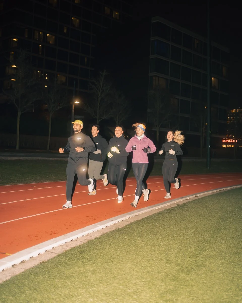 Group of five people running on an outdoor track at night, illuminated by a streetlight, with tall apartment buildings in the background.