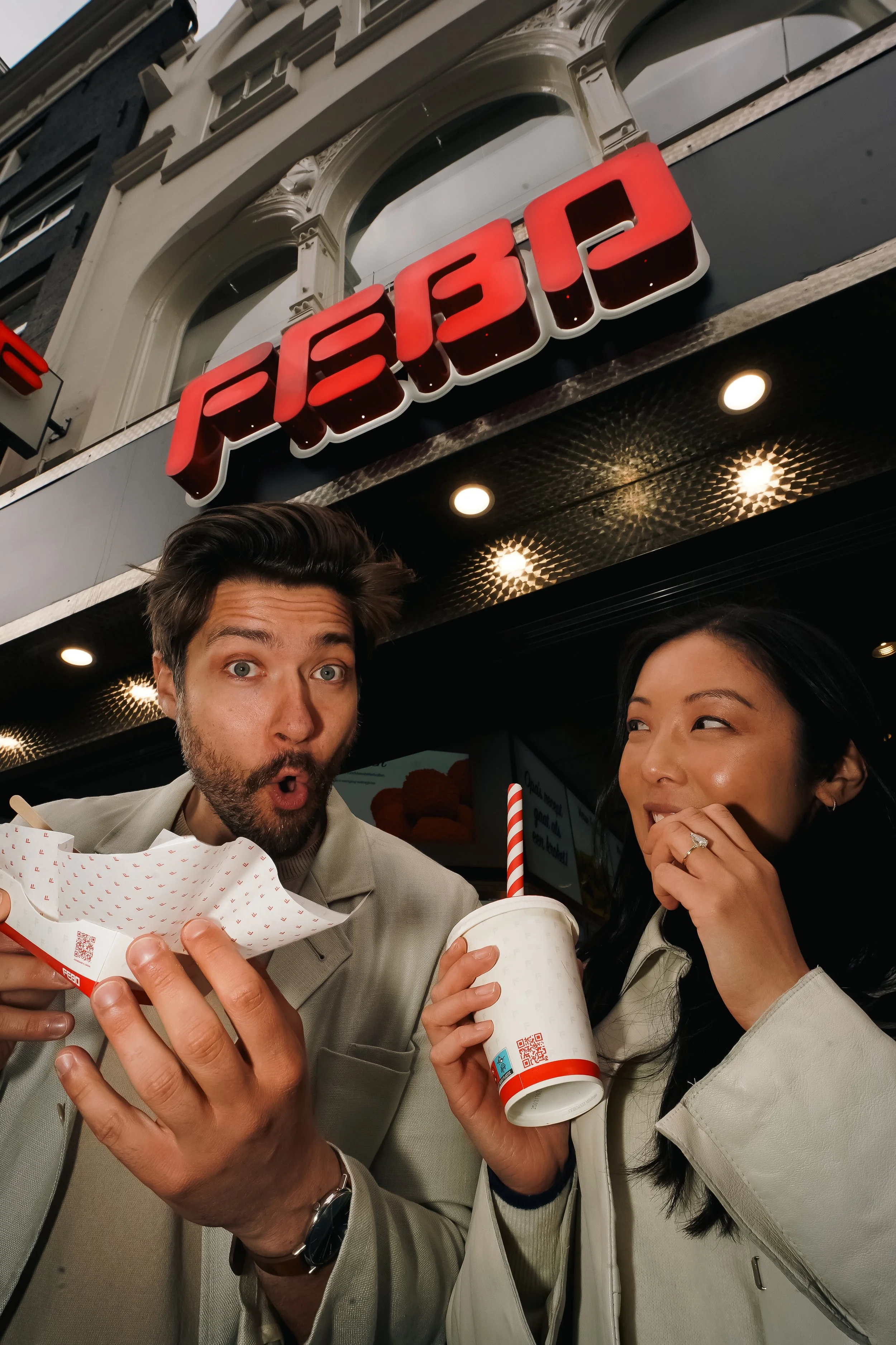 Two people enjoying fast food in a restaurant, with a man holding a paper food tray and a woman drinking from a large cup, standing in front of a store sign that reads 'PEIP.'