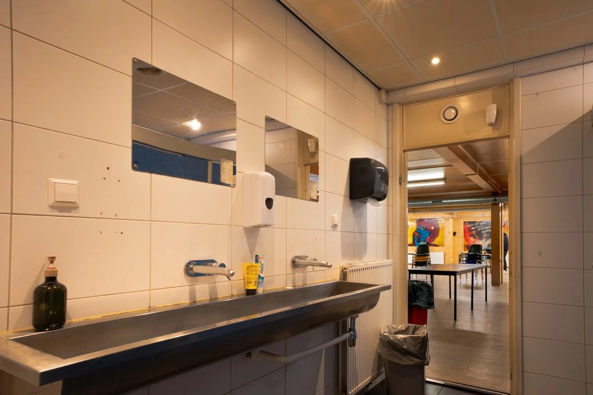 Public restroom with a stainless steel sink, soap dispenser, mirror, and paper towel dispenser, leading to a room with chairs and colorful wall art.