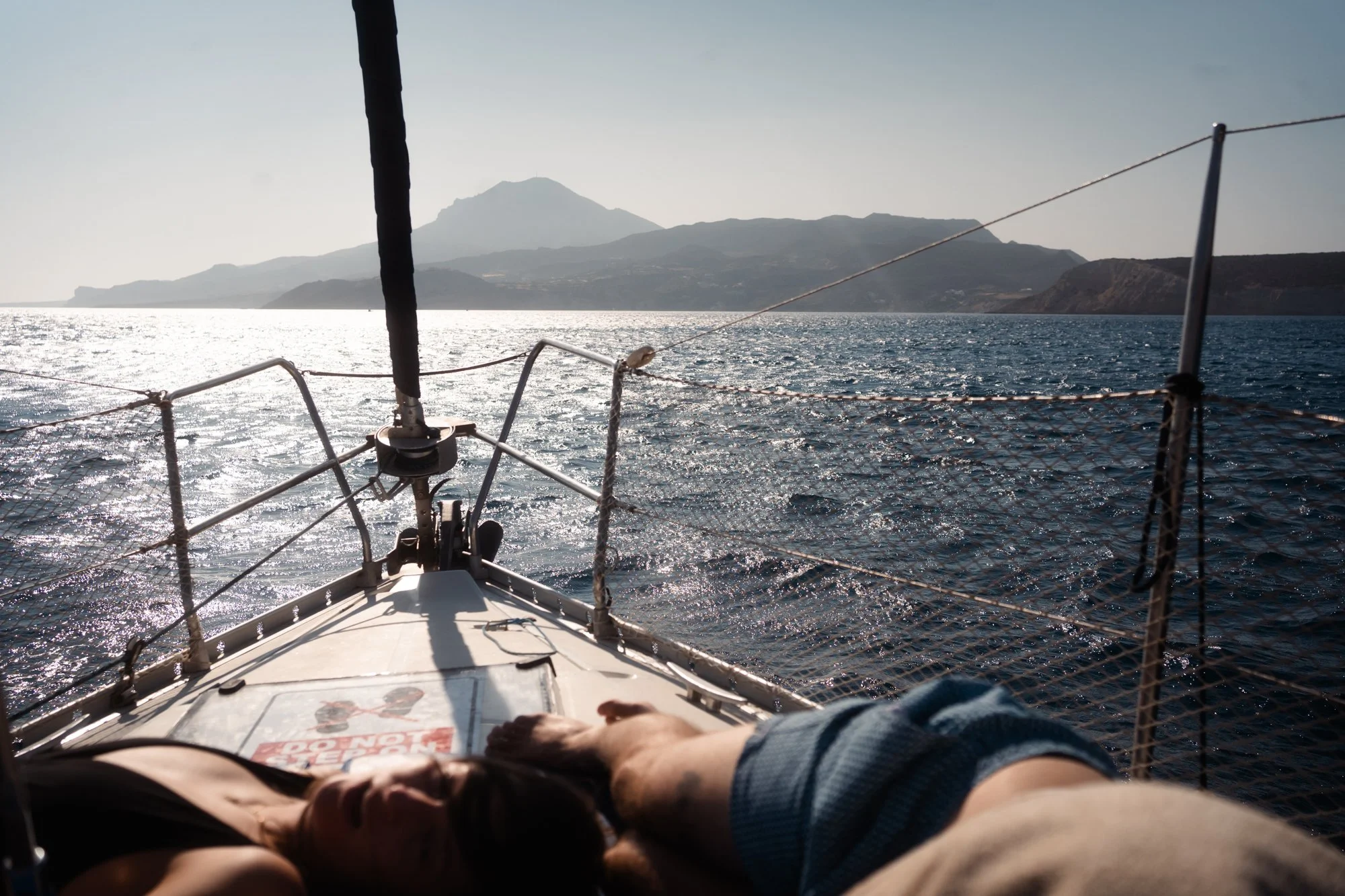 View from a sailboat showing a person’s feet and legs in the foreground, a white deck with some items, the ocean with shimmering water, and distant mountainous landmass with a prominent peak.