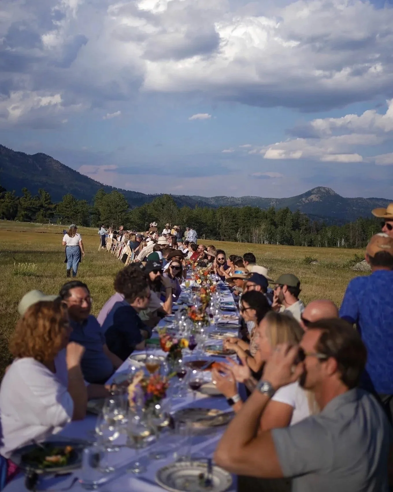 People are gathered outdoors for a long dining table set up in a field with mountains and clouds in the background.