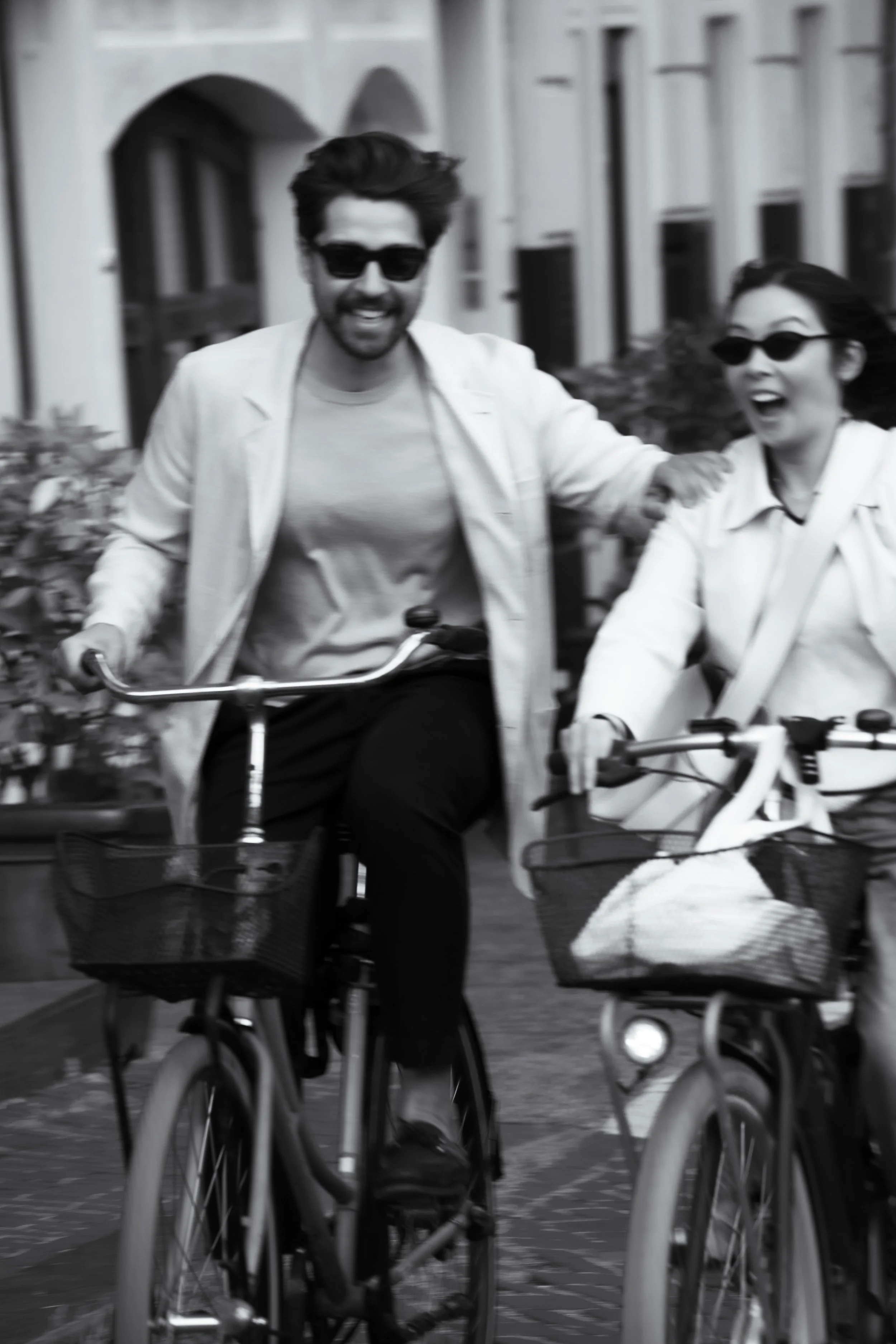 Two people riding bicycles outdoors, both wearing sunglasses, smiling and enjoying a sunny day.