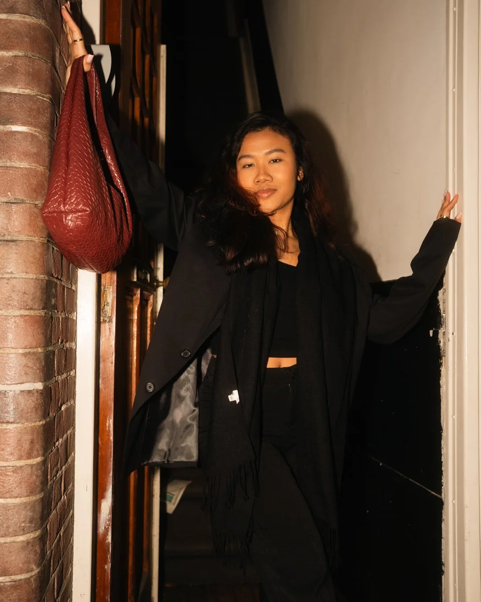 A woman with long dark hair wearing a black outfit and black blazer, standing in a doorway with one arm raised, holding a red handbag, with a brick wall on one side.