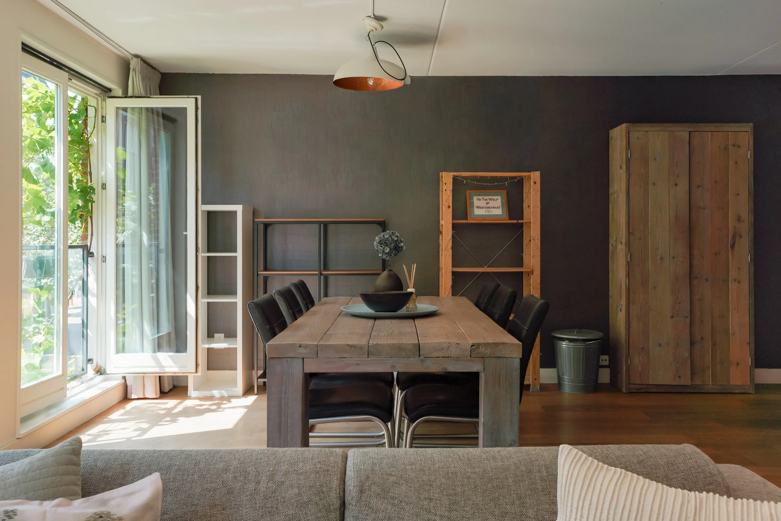 A dining area with a wooden table and black chairs, next to a large open window with curtains, a dark accent wall, shelves, framed artwork, and a wooden cabinet, with sunlight streaming in.